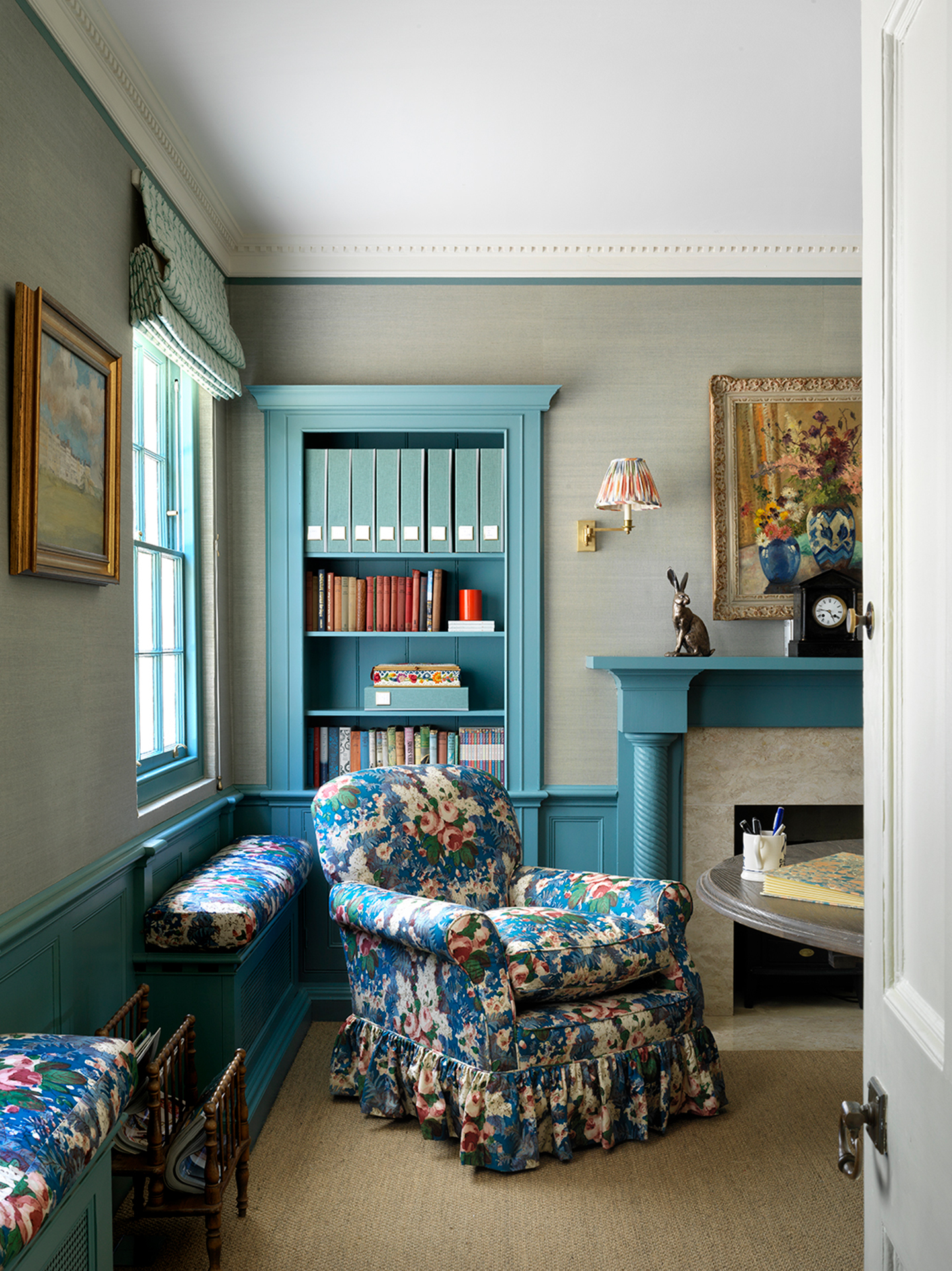 An office with textured light green wallpaper on the upper half of the walls and painted blue panelling on the lower half. A floral frilled armchair sits in front of a built-in bookcase.