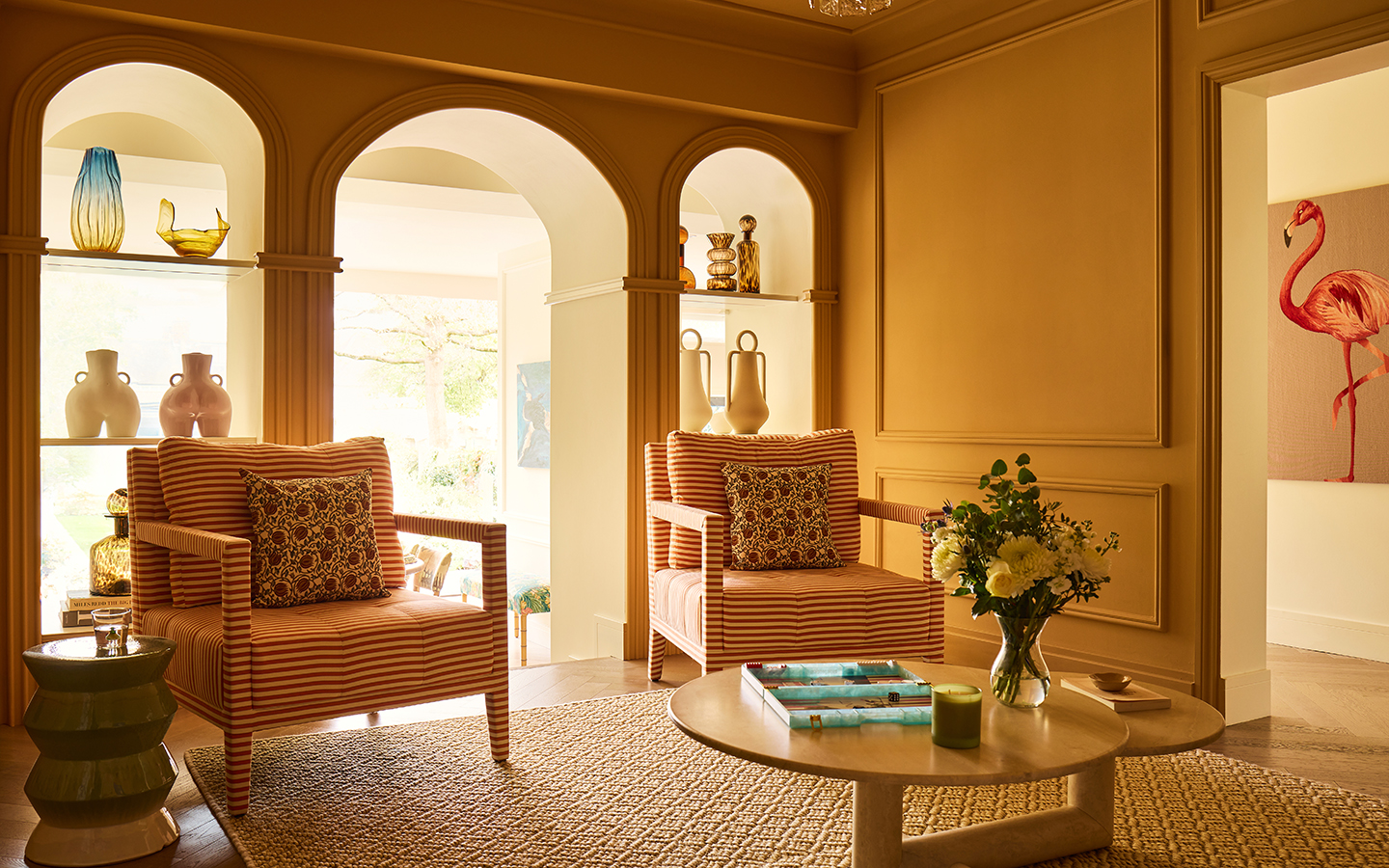 A seating area with golden yellow walls. The back wall feature three arches, with the two side arches featuring shelves lined with vases. A pair of striped armchairs sits in front of the arches, on top of a carpet and next to a wooden coffee table