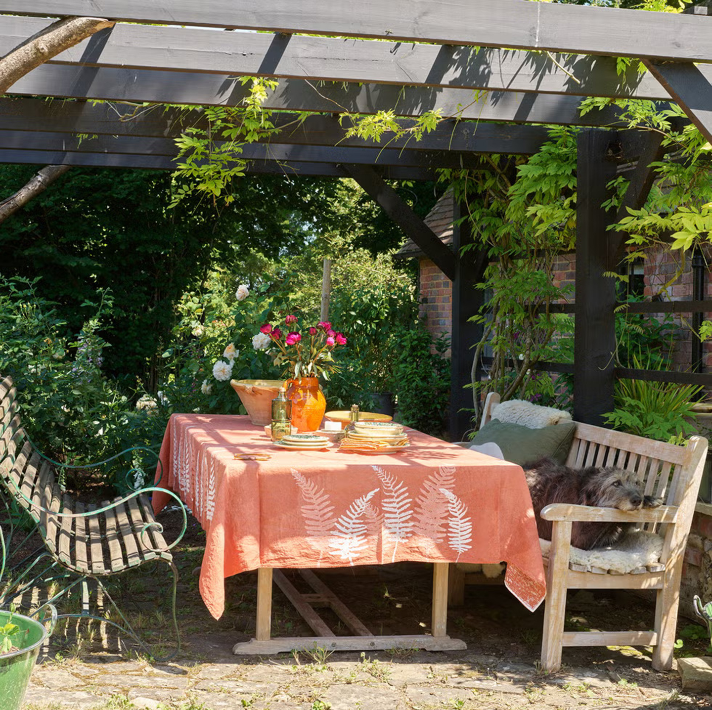 a garden with a wooden pergola with a dining table covered in an orange tablecloth underneath and a lurcher laid on the bench