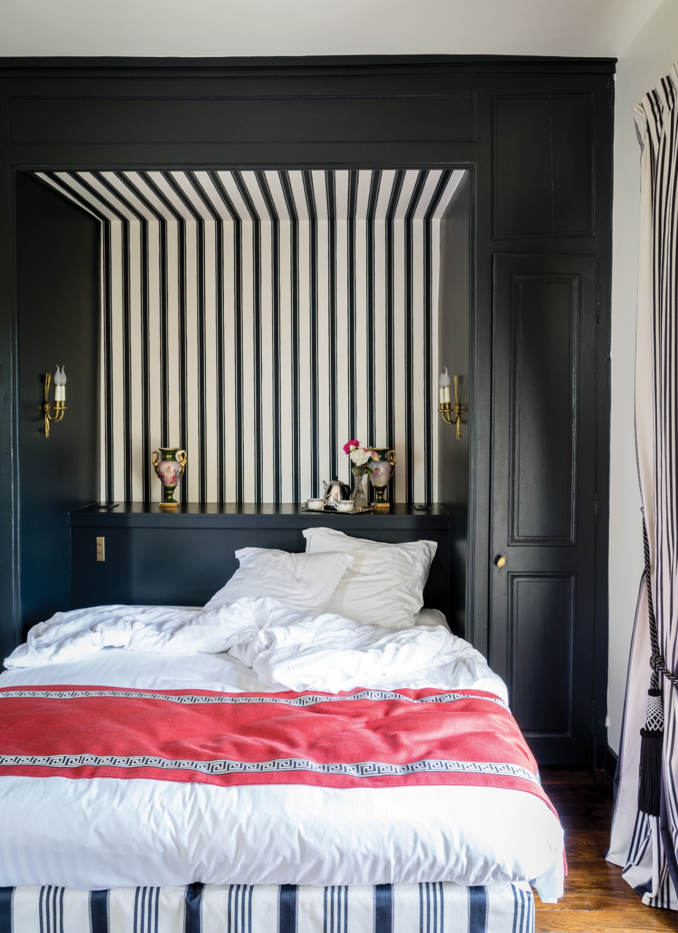 Black built-in bed nook featuring bold black-and-white vertical striped wallpaper inside the alcove, brass wall sconces, white bedding, and a coral throw blanket