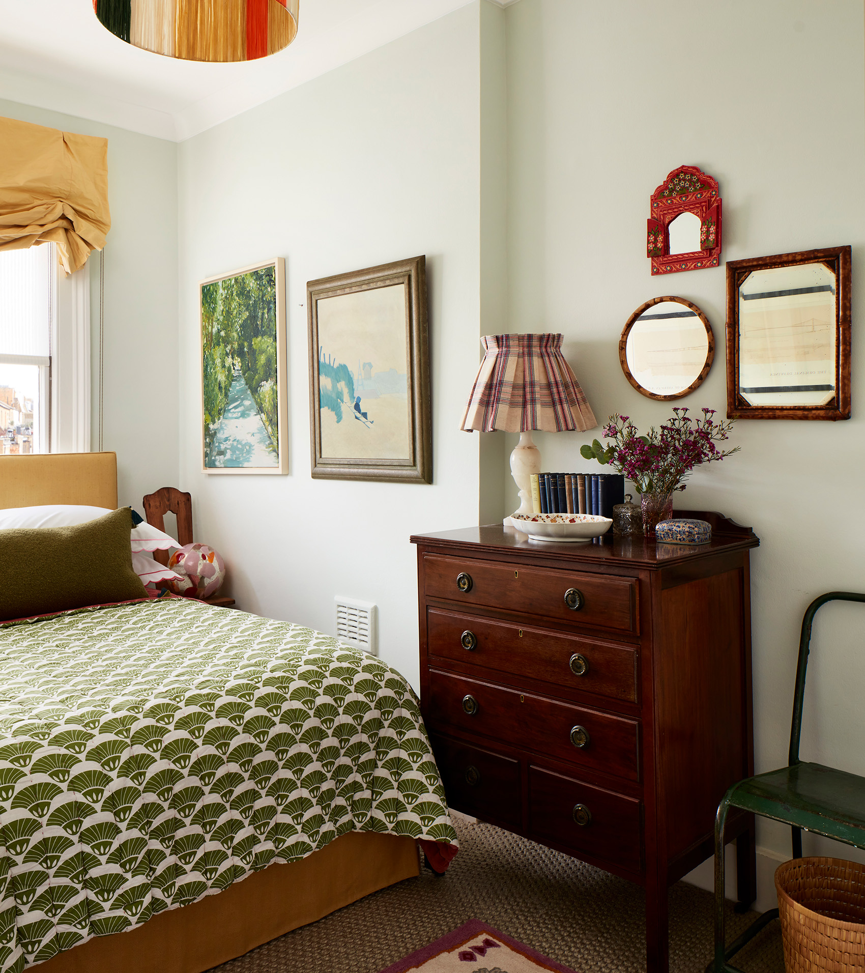 a bedroom with pale walls, a mahogany chest of drawers, art and mirrors hung on the wall and a bed with a green and white patterned throw