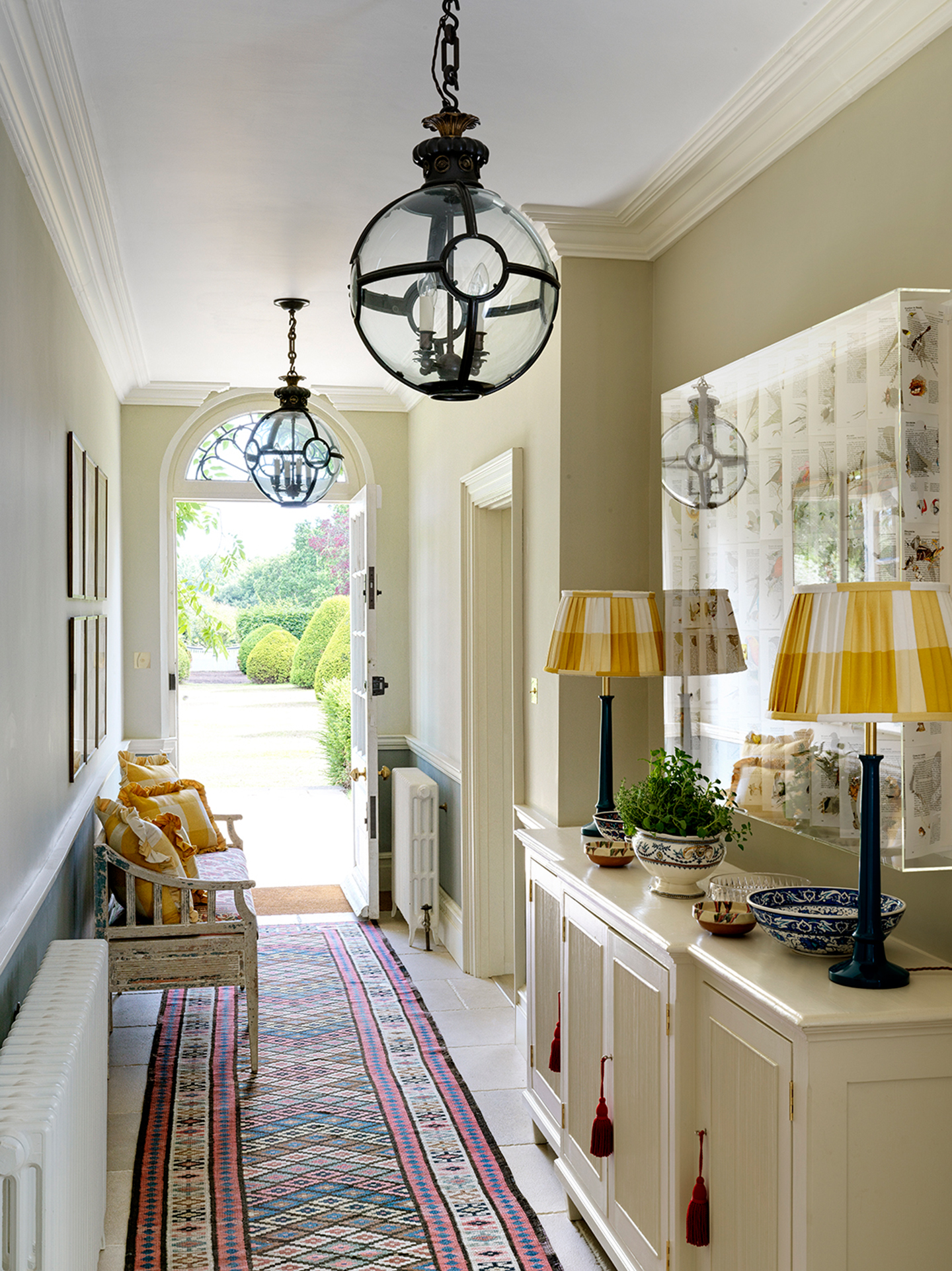 A hallway with an open front door at the end. A colourful runner runs along the floor and two large globe lanterns hang above. A bench sits to the left and a sideboard to the right, topped with table lamps and ceramics. A large display case hangs above