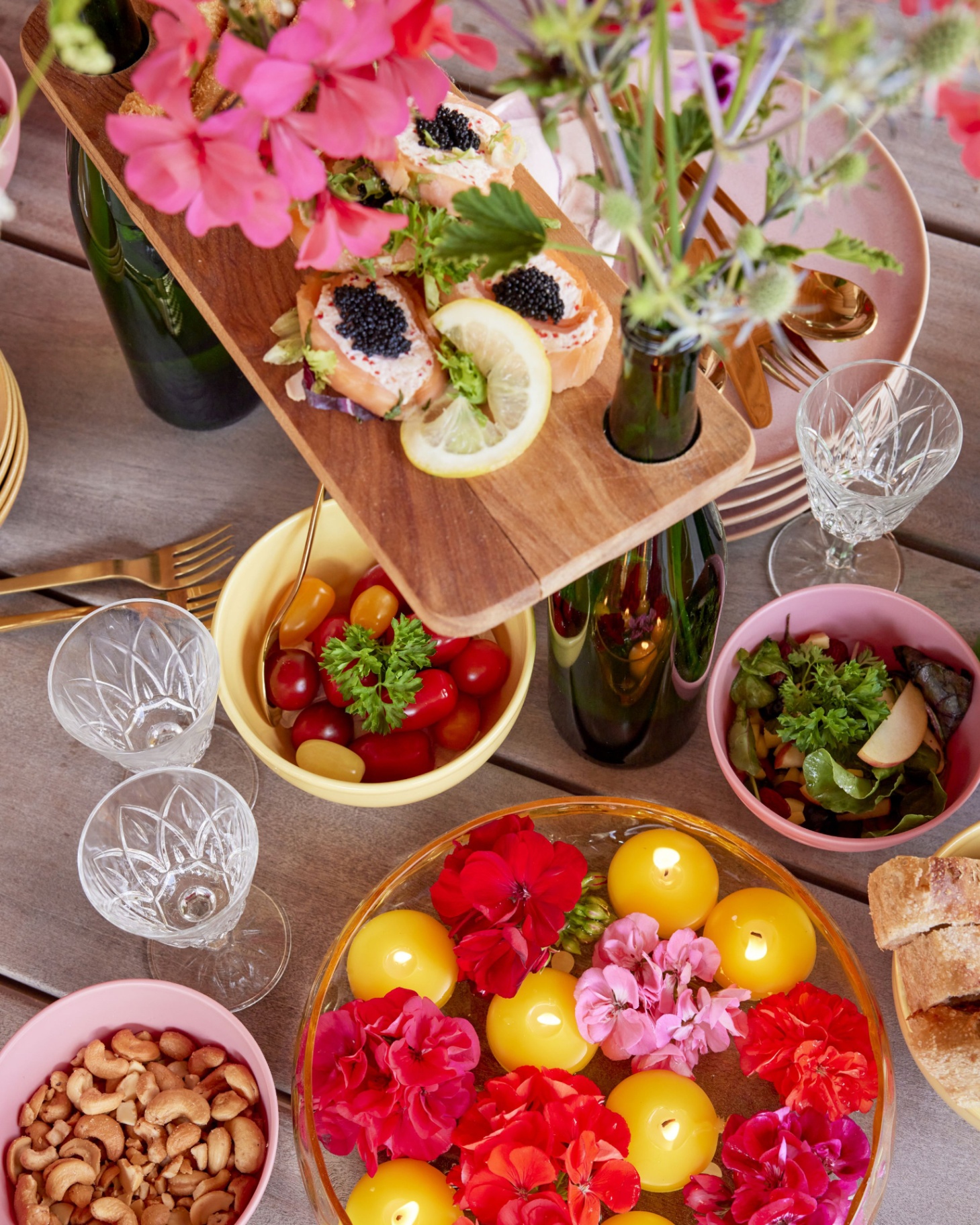a table filled with party food, a bowl of cashews, a bowl of tomatoes, a rasied wooden board with bread and caviar and pink and red floral decorations