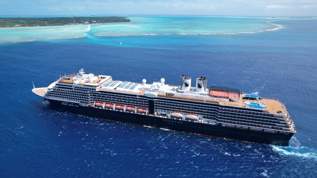 High-angle aerial view of the Holland America Line cruise ship Zuiderdam cruising through deep blue tropical ocean waters near a shallow turquoise reef and island shoreline