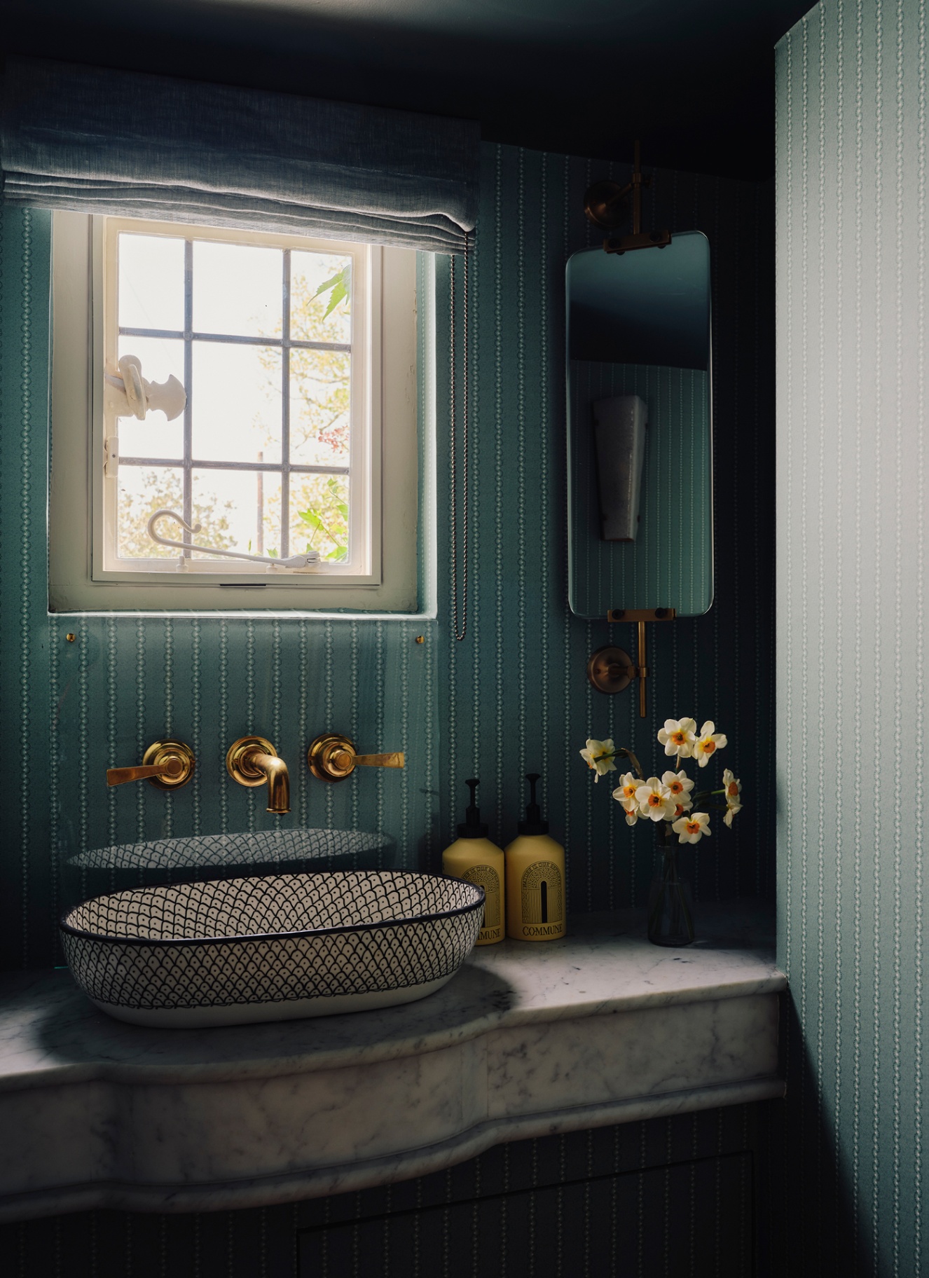a downstairs toilet with teal striped wallpaper, a marble countertop with a black and white patterned basin, a corner mirror and a window with a blue blind and a vase of daffodils