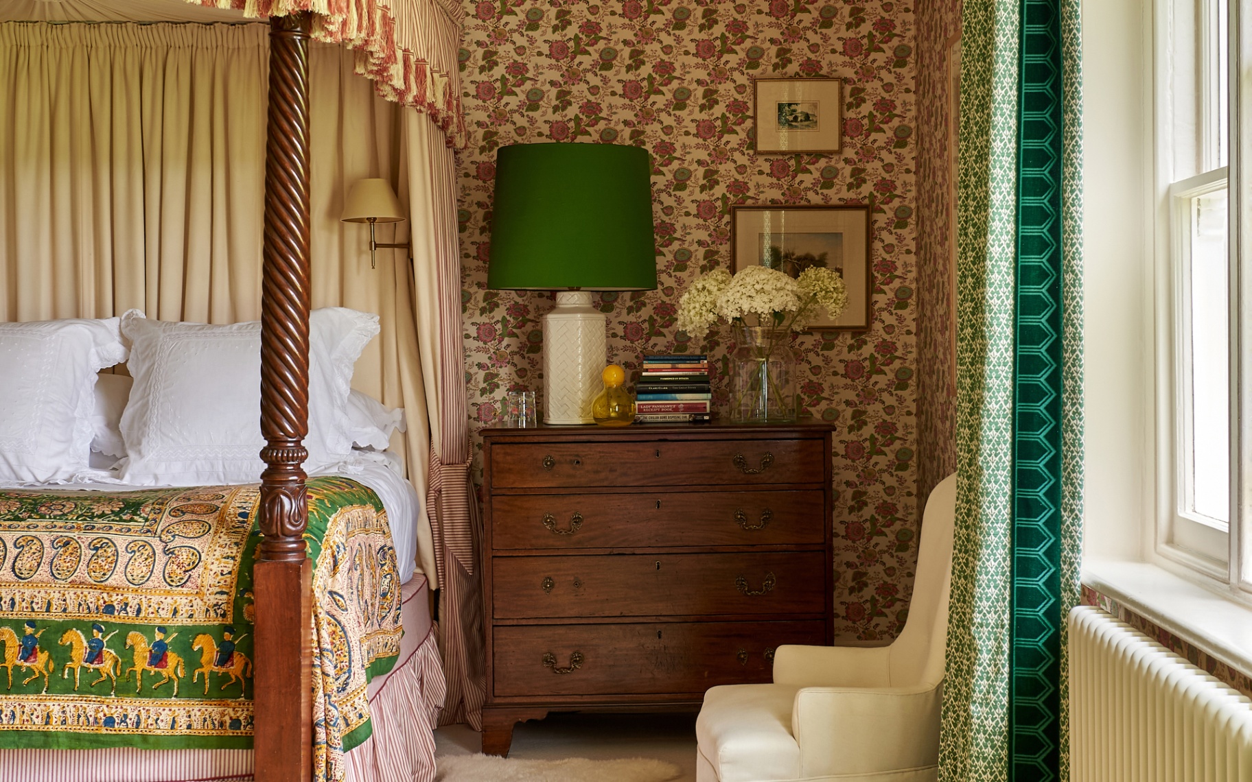 a bedroom with a pink and brown floral wallpaper, an antique wooden chest of drawers with a green lamp, a cream accent chair and a large wooden four poster bed with a green and yellow patterned quilt