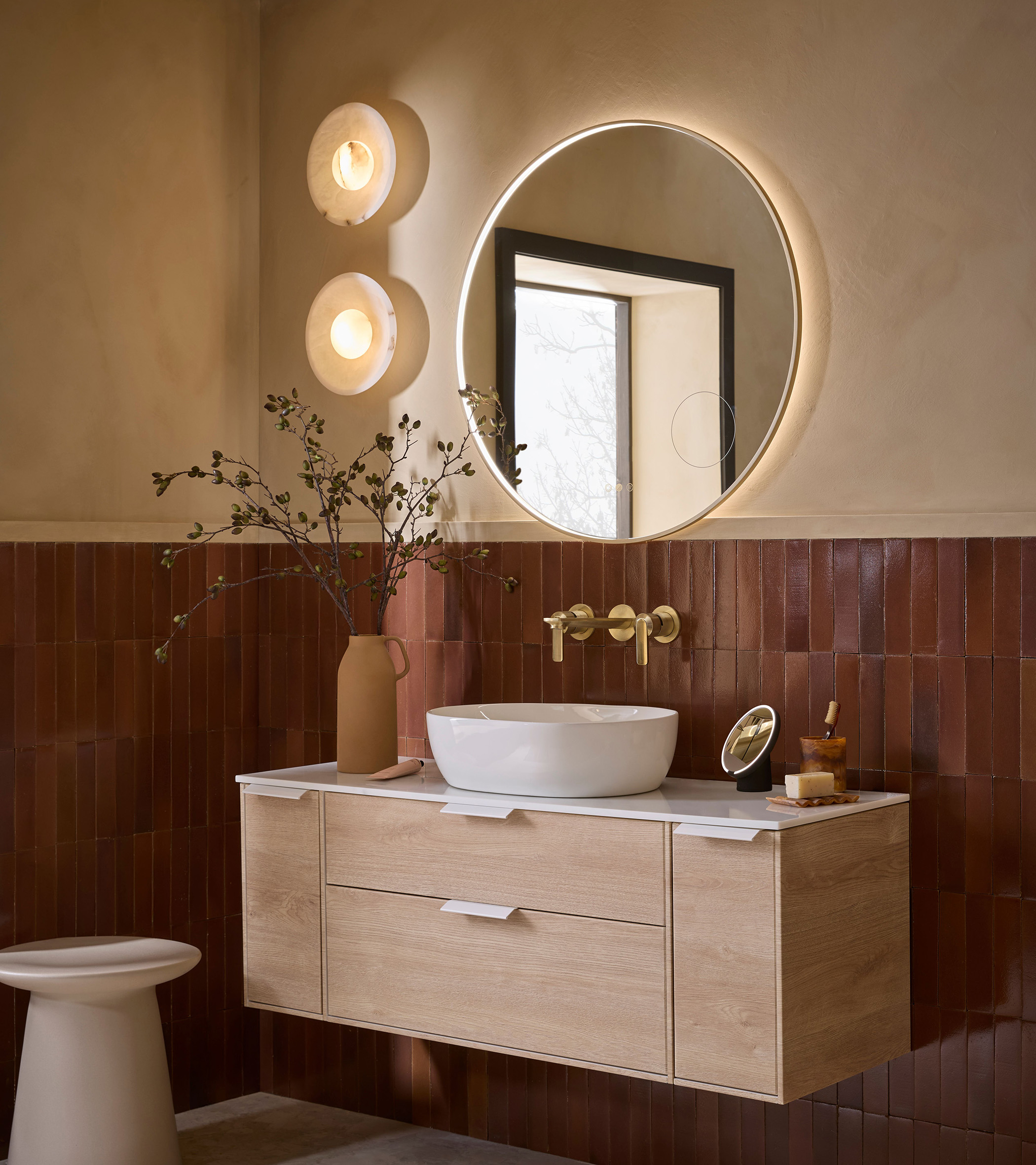 a bathroom corner with half brown tiled walls and half cream, a floating light wood vanity with a circular light up mirror hanging above it