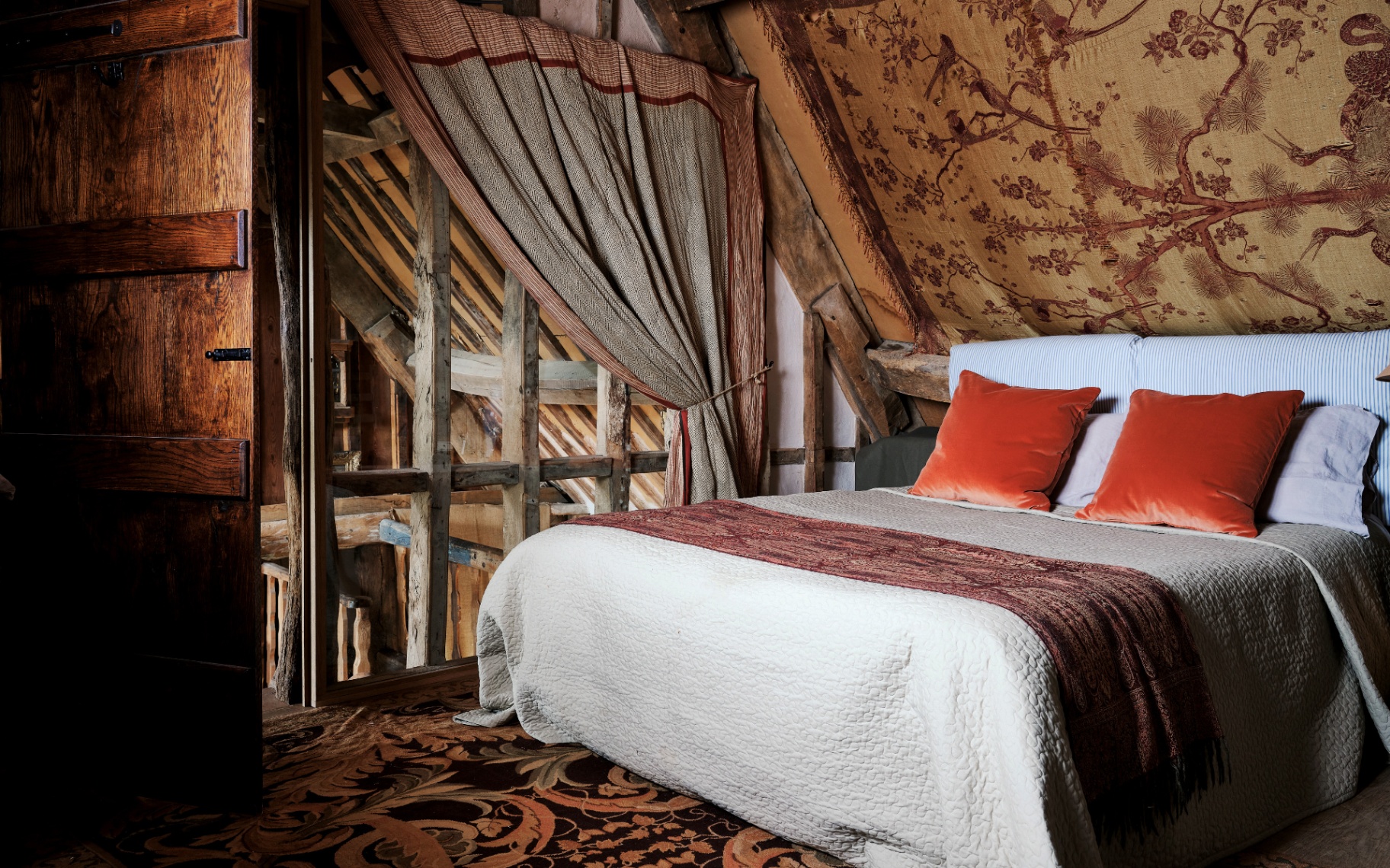 Rustic attic bedroom under timber beams featuring a toile-patterned sloping ceiling, cream bedspread with burnt orange pillows, patterned carpet, and exposed wooden frame