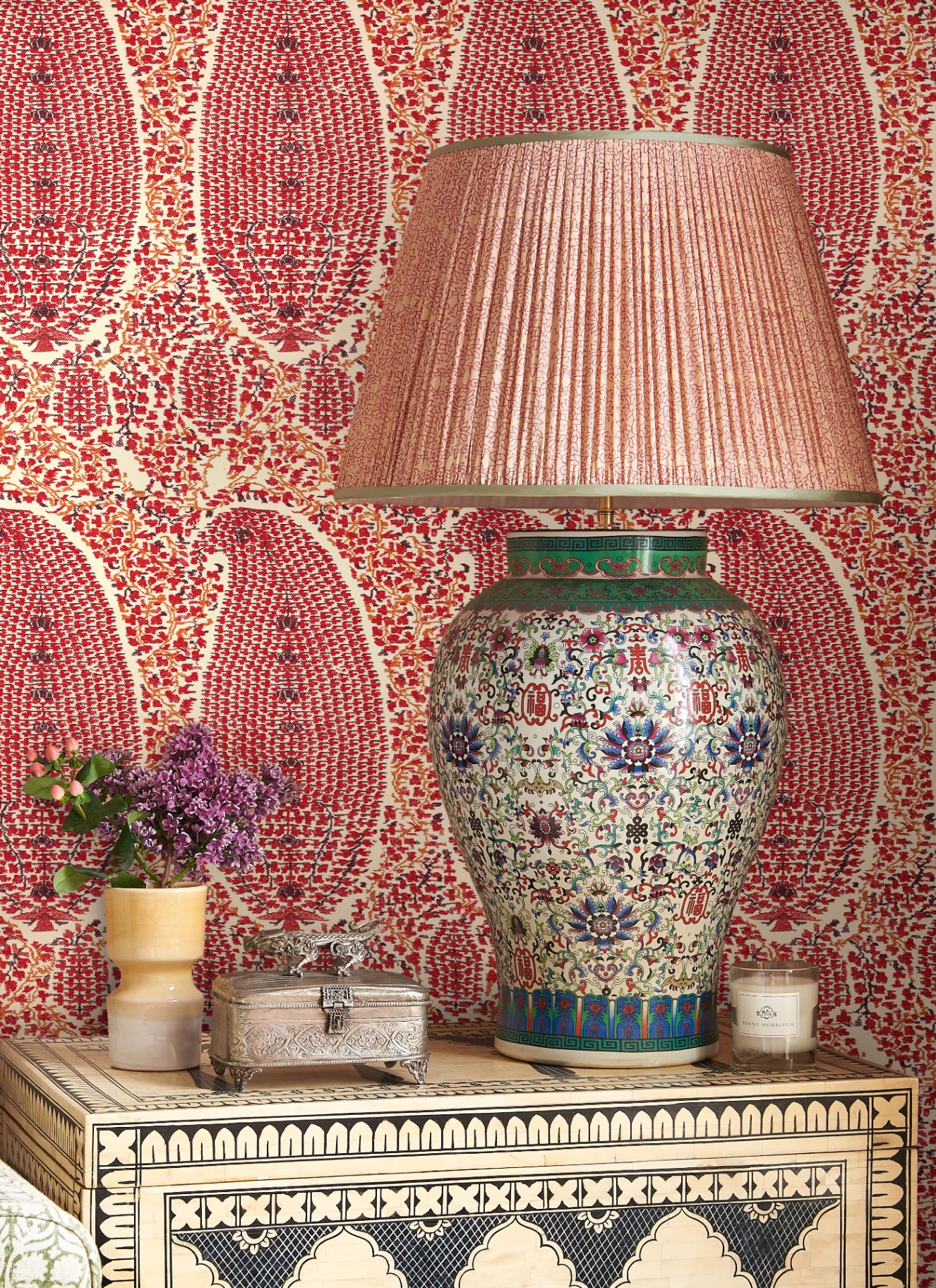 a close up of a wooden sideboard painted with black inticate patterns, a red and white botanical wallpaper and a large ceramic blue and red patterned lamp base with a pale pink pleated shade