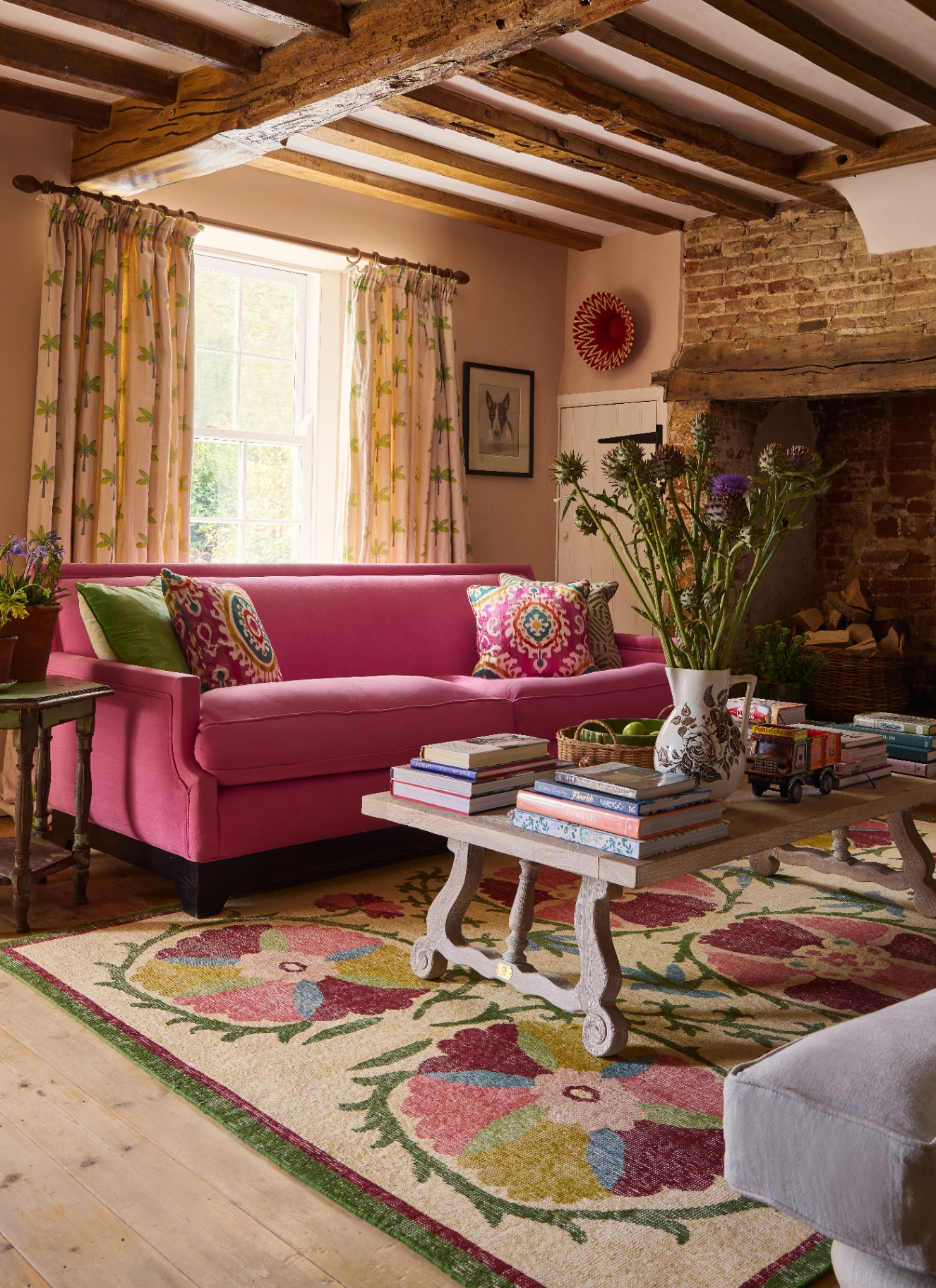 A bright pink sofa with patterned cushions sits on a bold floral rug in a rustic living room with exposed wood beams, a brick fireplace, palm-tree curtains, and a book-stacked coffee table