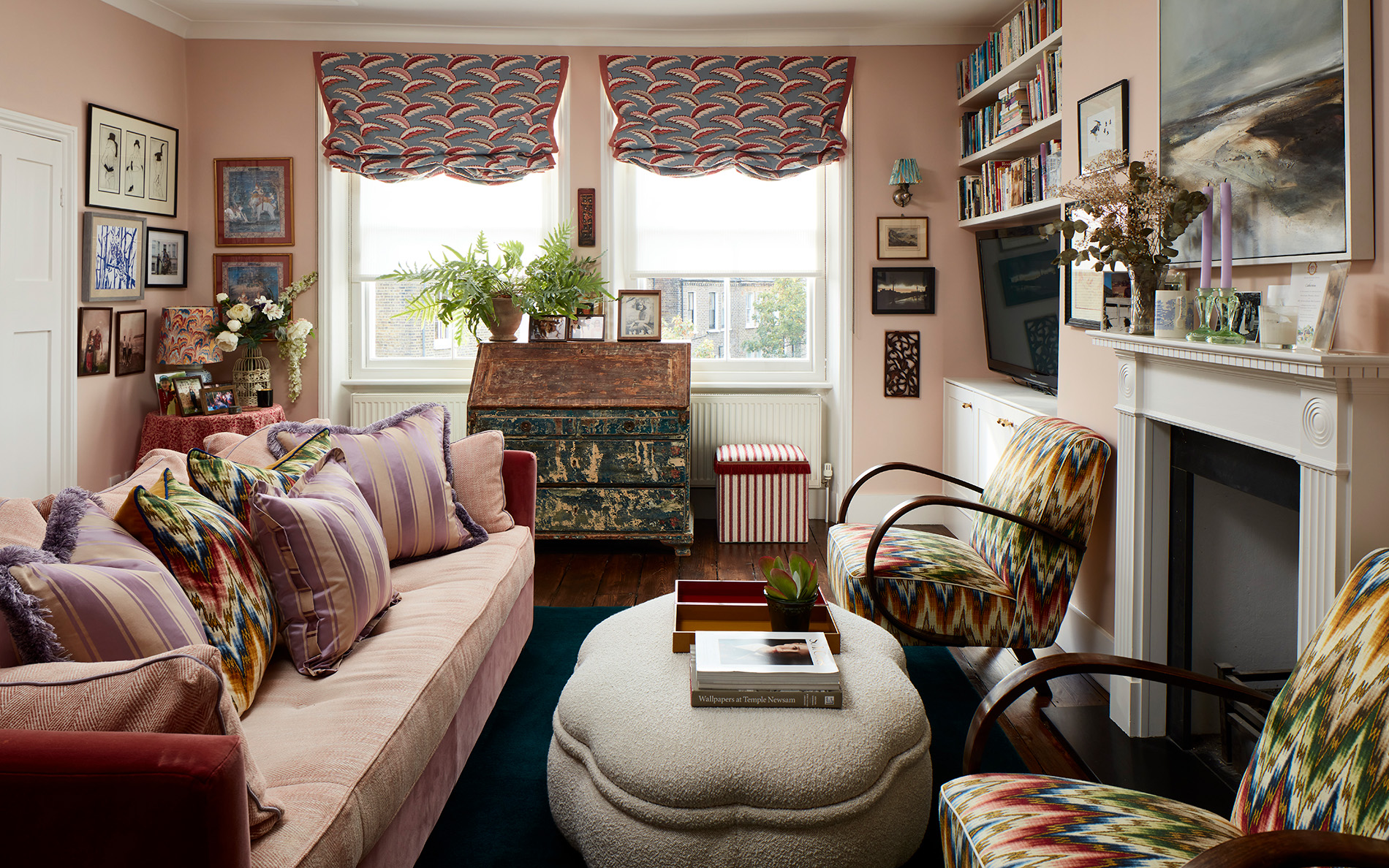 a small living room with pale pink walls, a pale pink sofa with patterned cushions on the left, two colourful patterned armchairs on the right in front of a fireplace and a round borg ottoman in the middle