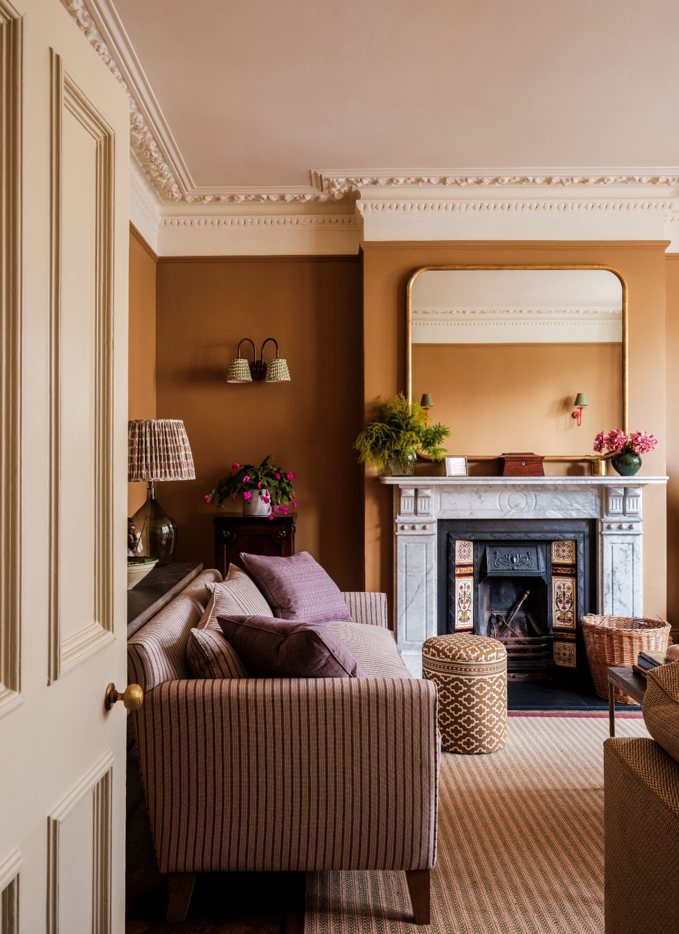 A warm, ochre-painted living room featuring a marble fireplace with a large gilt mirror, a striped sofa with mauve cushions, a patterned ottoman, and detailed ceiling cornicing.