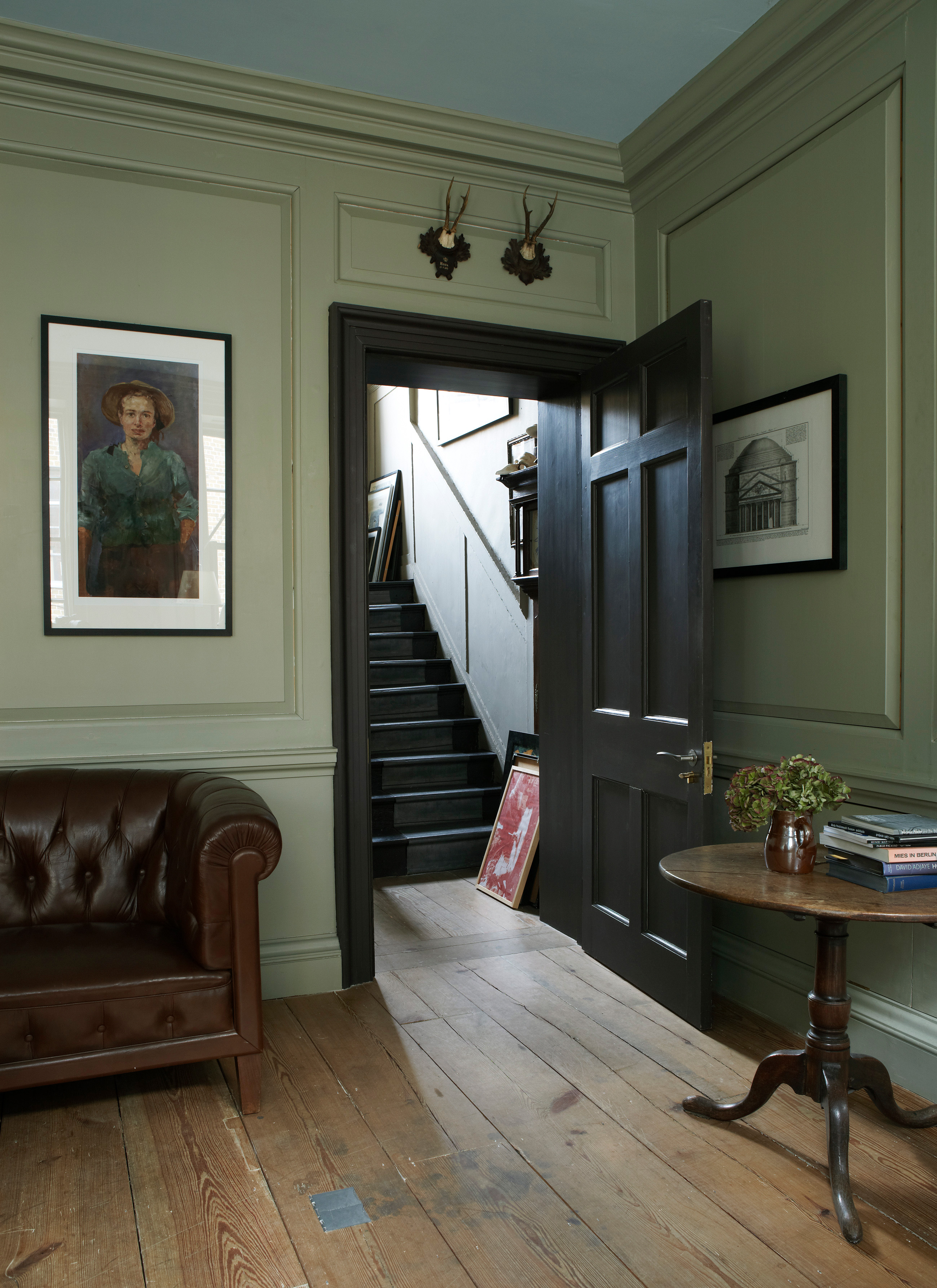 a living room with wooden flooring, pale green walls, a large black open door and a portrait hanging next to it