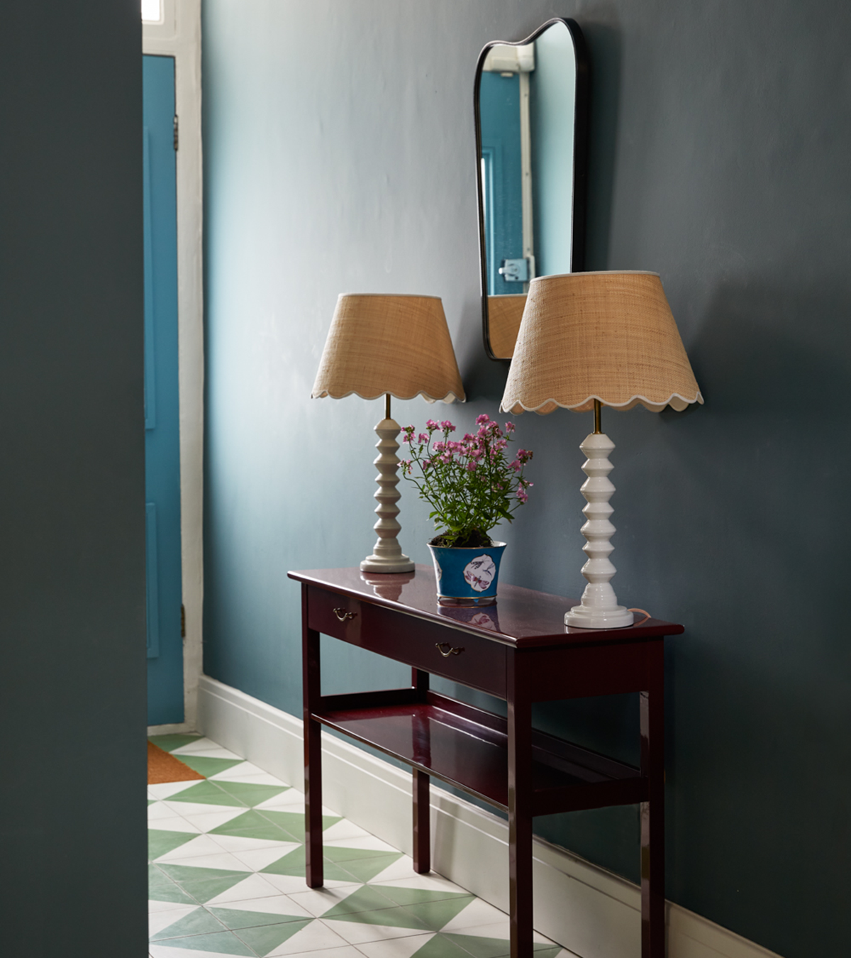 a hallway with dark navy walls, checkered floor, a mahogany console table with two large white lamps and a large mirror above
