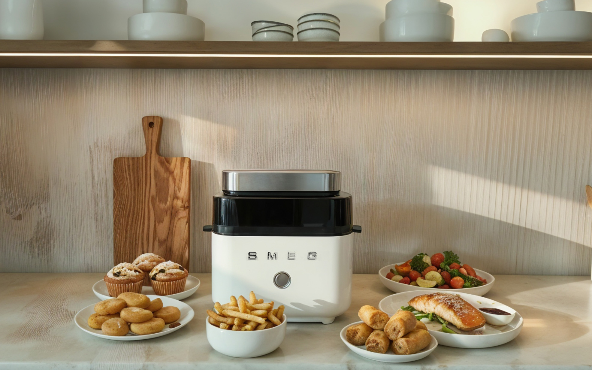 A white SMEG air fryer sitting on a modern kitchen counter surrounded by plates of cooked food, including french fries, muffins, spring rolls, baked salmon, and a fresh salad. A wooden cutting board leans against the wall in the background