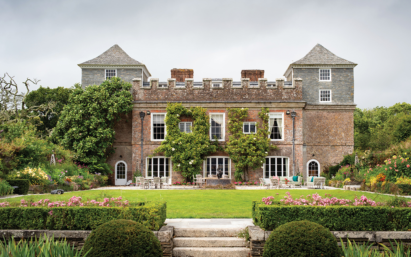 a straight on shot of a 17th century manor house with battlemented towers on either end and a neat lawn and gardens in front