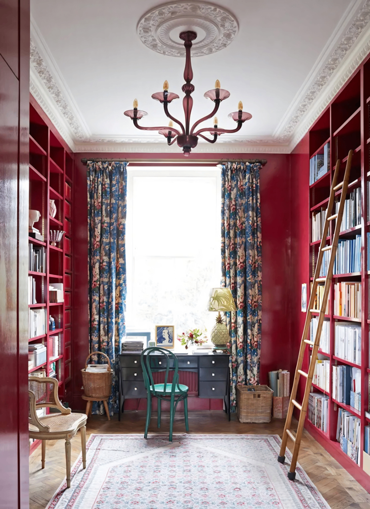 Home office with custom floor-to-ceiling bookshelves, walls, and window frames painted in a high-gloss crimson red, featuring a traditional chandelier, green chair, and patterned curtains
