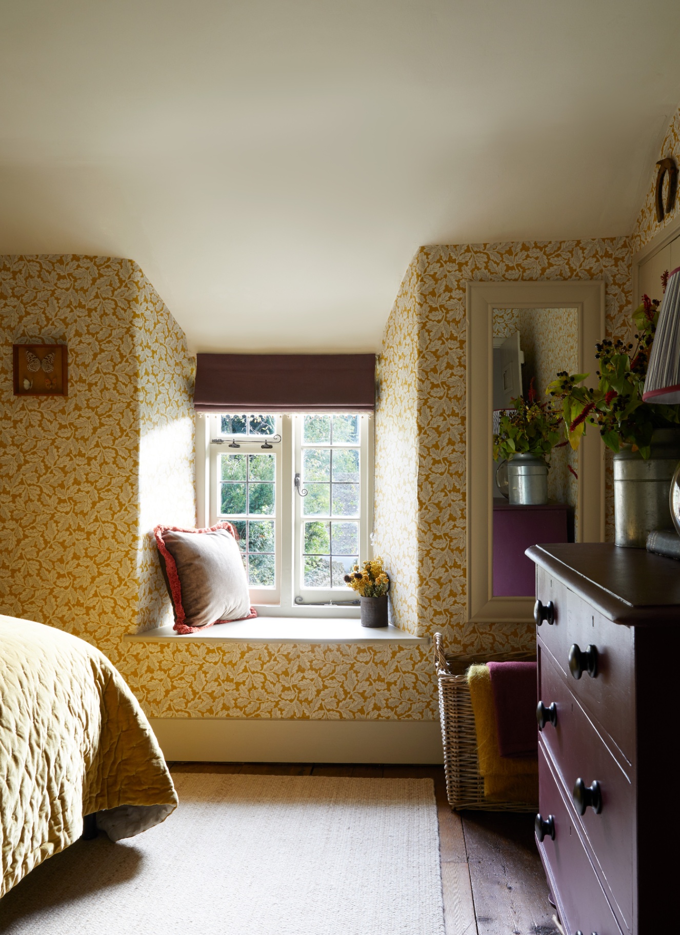 a bedroom with yellow and white botanical wallpaper, a large dark purple chest of drawers and a window seat nook