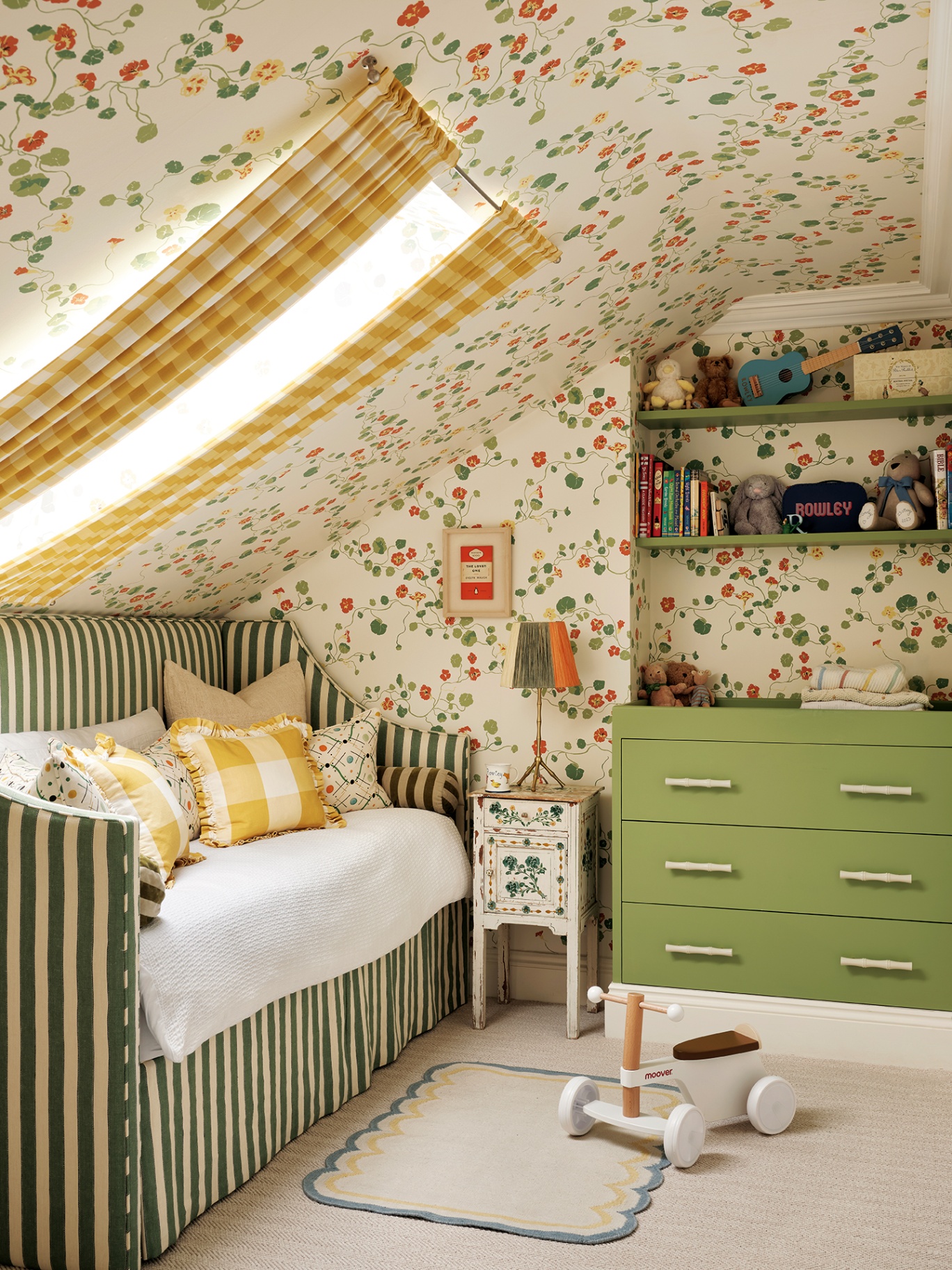 a playful nursery with a sloped ceiling above a striped green and white daybed dressed with frilled and patterned cushions. The walls and ceiling are papered in a botanical wallpaper and at the back of the room is a green chest of drawers