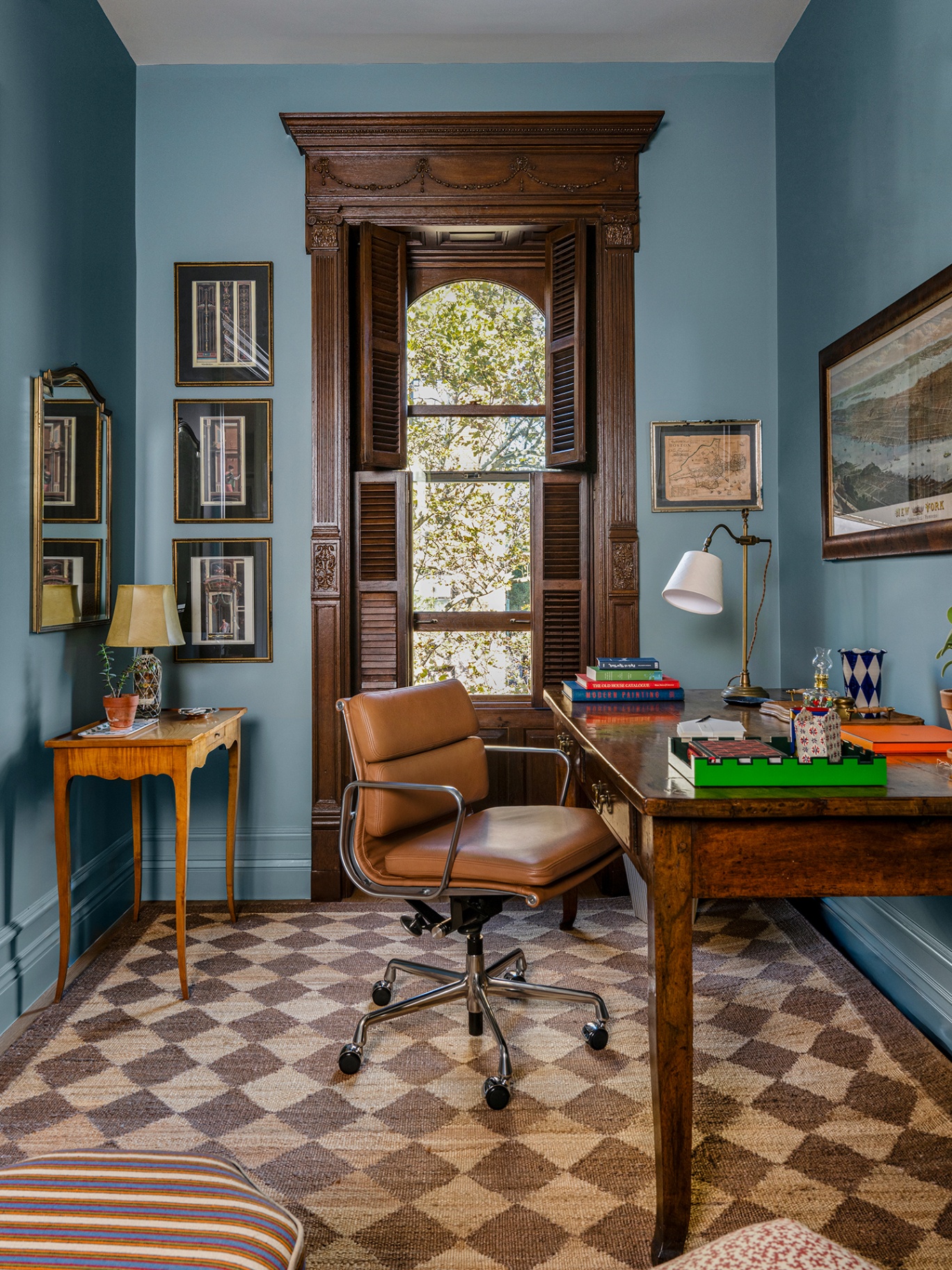 A home office with blue walls and a large dark wooden window with pediment on the far wall. A wooden desk and leather swivel chair sit on a diamond pattern rug. To the left is a small console table. Architectural and geographical prints hang on the walls 