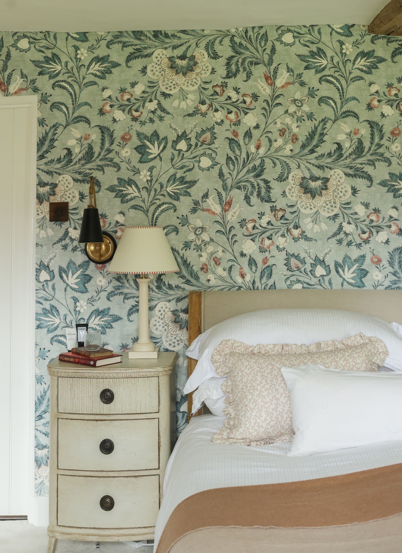 a bedroom with pale green botanical wallpaper, a bed with white bedding and ochre throw and a shabby chic bedside table