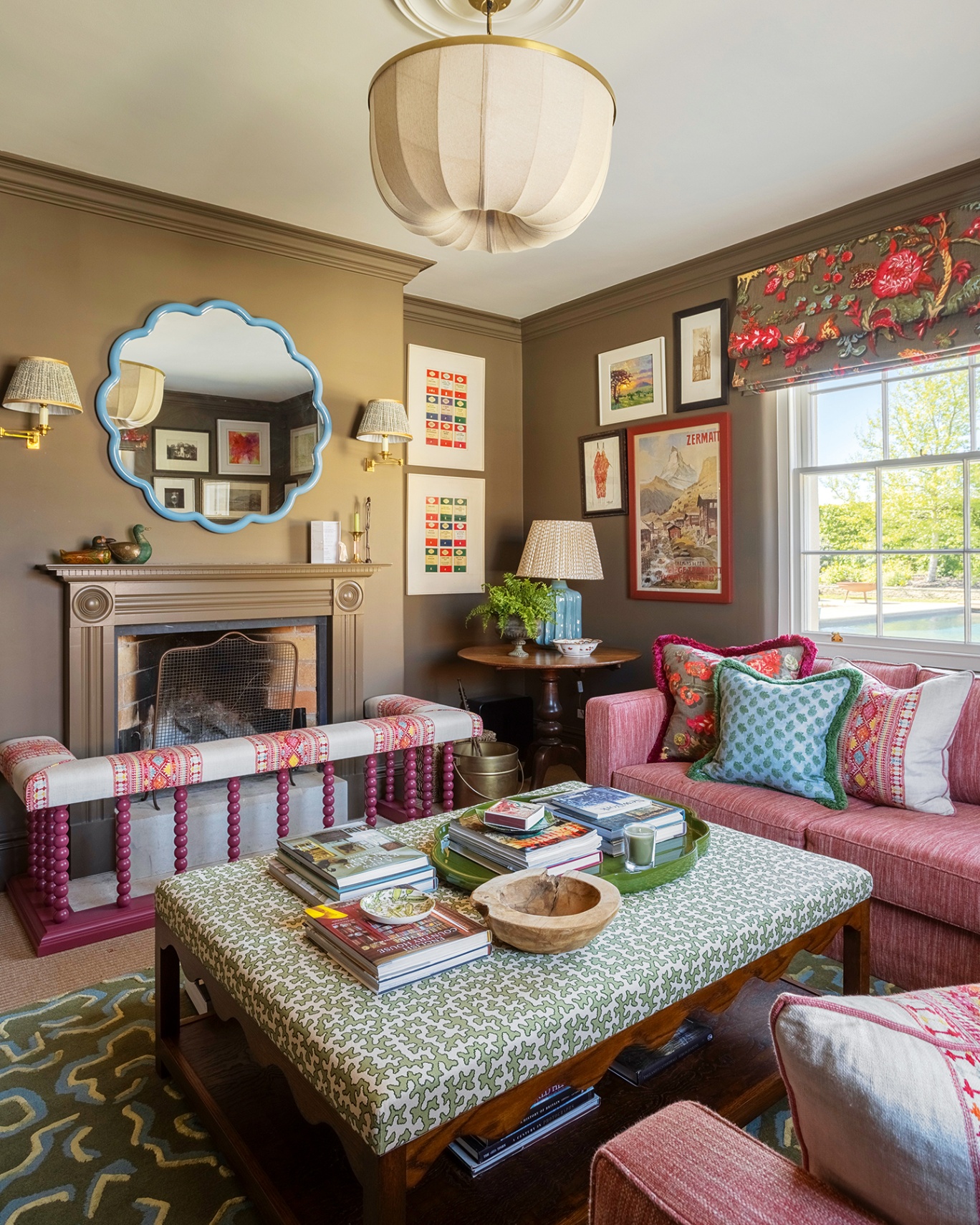 A dark green snug with a large upholstered ottoman in the foreground, surrounded by two pink sofas dressed with decorative cushions. A large fabric pendant hangs above and a fireplace is paired with a patterned bobbin fender and a blue overmantel mirror