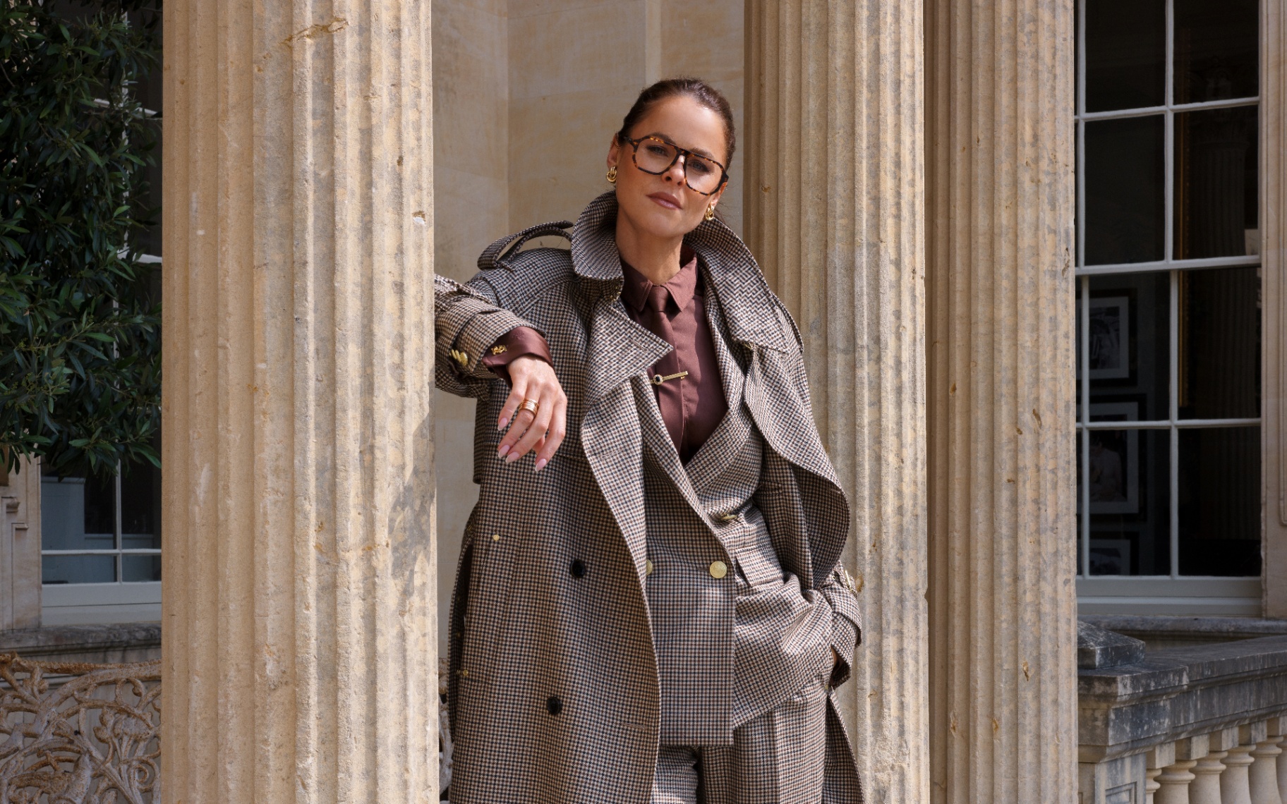 Jade Holland Cooper wearing a tailored houndstooth trench coat and matching suit, leaning against stone columns outside her country home