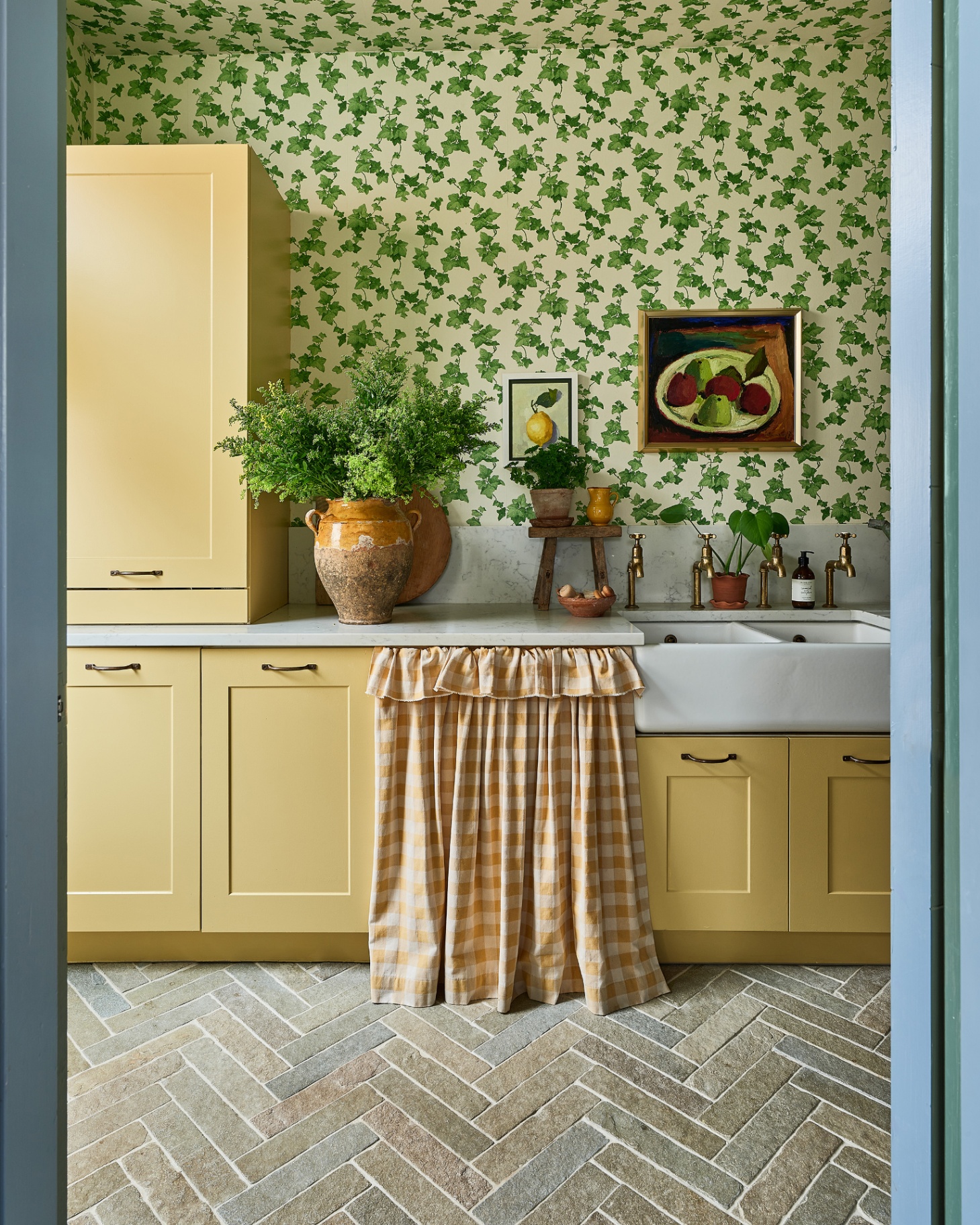 a utility room with green ivy trailing wallpaper, yellow cabinets, a yellow and white gingham fabric skirt covering a cupboard and grey stone tiled floor