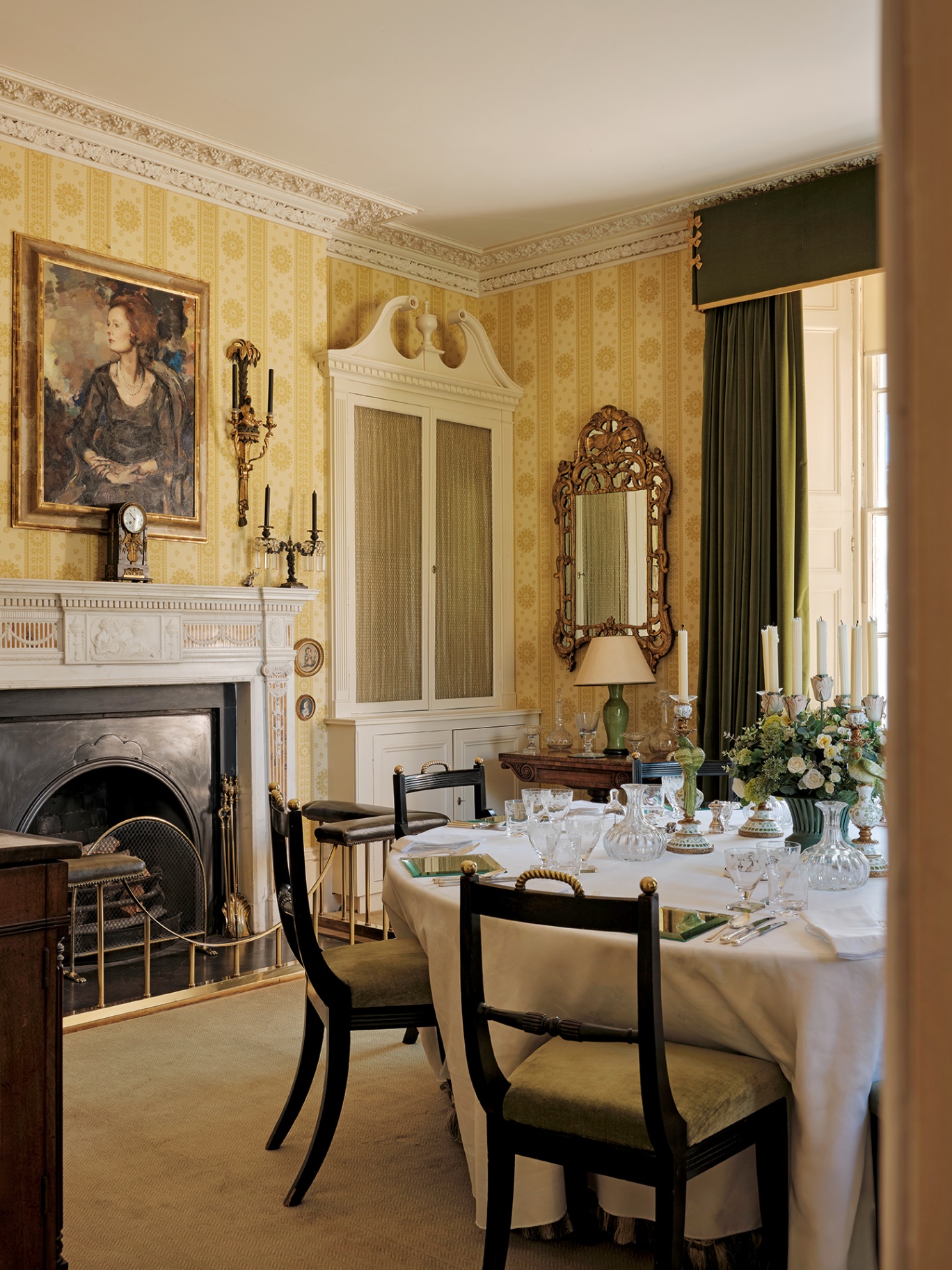 a classic English country dining room with yellow striped wallpaper, an ornate fireplace, built-in cabinet with an intricate pediment and a large circular table surrounded by wooden chairs and laid with a white cloth and pretty glassware