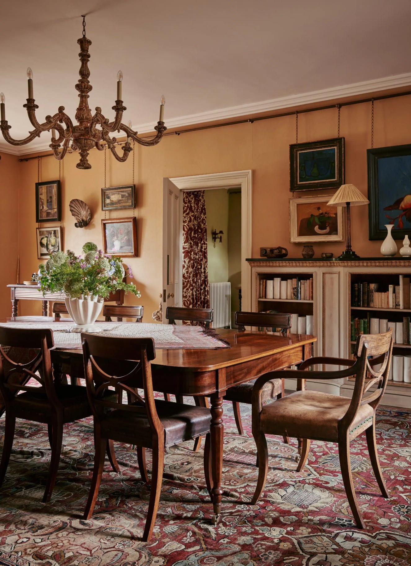 Warm terracotta dining room with traditional carved wooden dining chairs, a large dining table over an intricate patterned Persian rug, classic chandelier, and art hung on picture rails