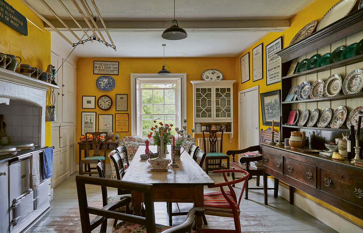 a bright yellow kitchen with a central wooden dining table and chairs. To the right is a large wooden dresser filled with plates and to the left is an Aga with a hanging clothes airer above
