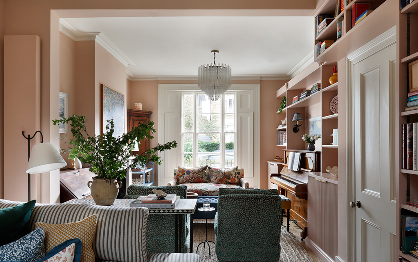 an elegant living room with plaster pink walls, a large white chandelier, a wooden piano, open shelving and a floral sofa and a stripe sofa