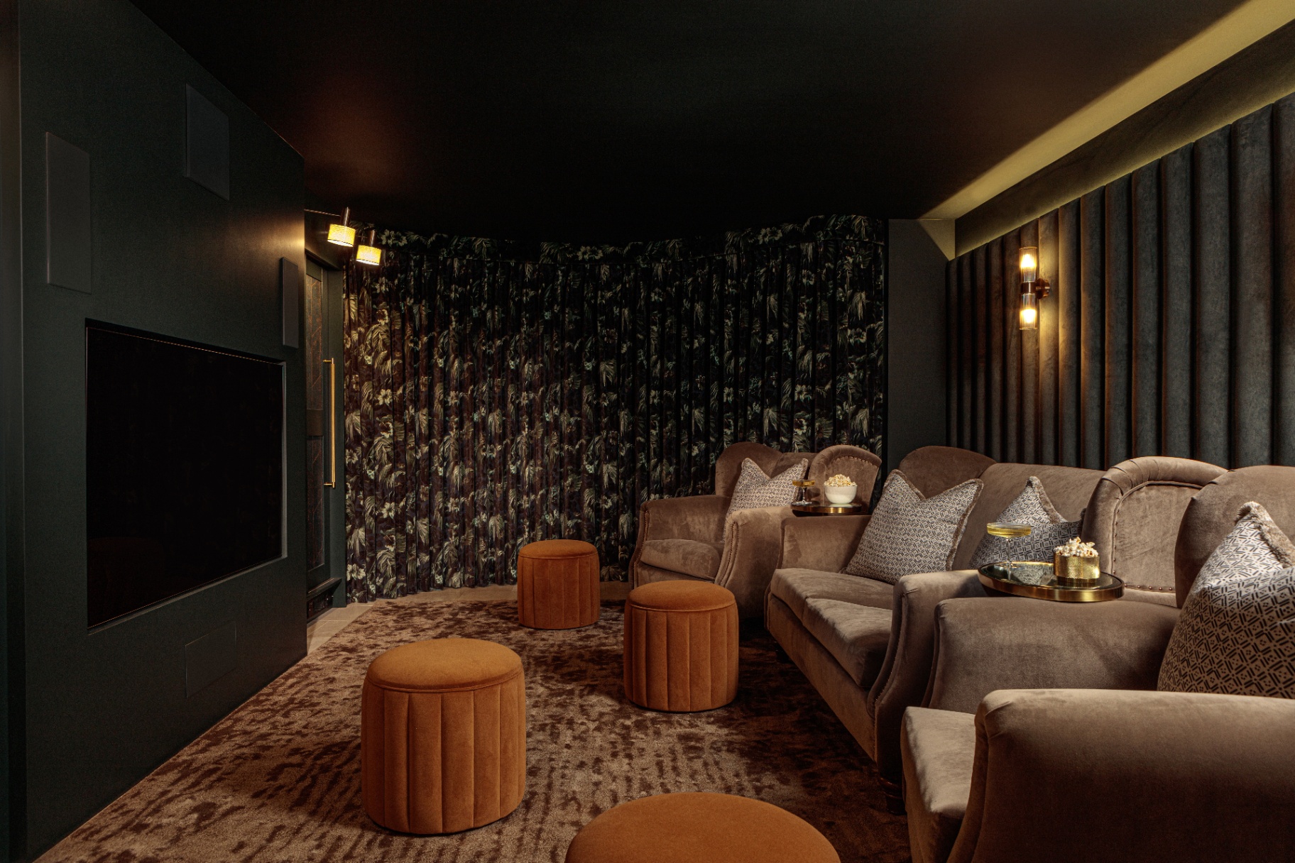 an indoor home cinema room with dark floral wallpaper, a large screen and grey velvet armchairs and rust orange velvet footstools