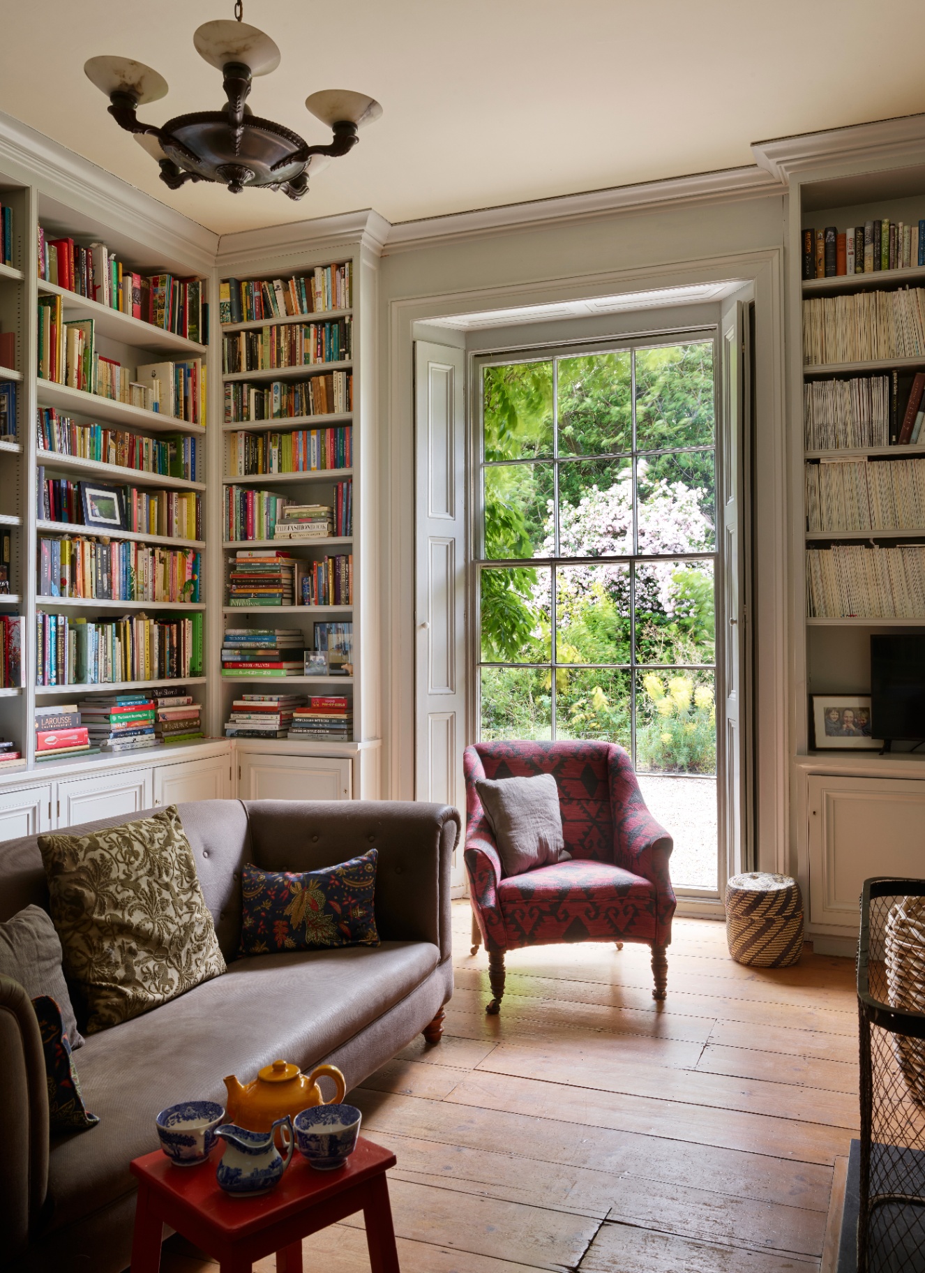 A light-filled room with floor-to-ceiling bookshelves surrounding a tall sash window with garden views, accompanied by a patterned pink armchair and a low grey sofa