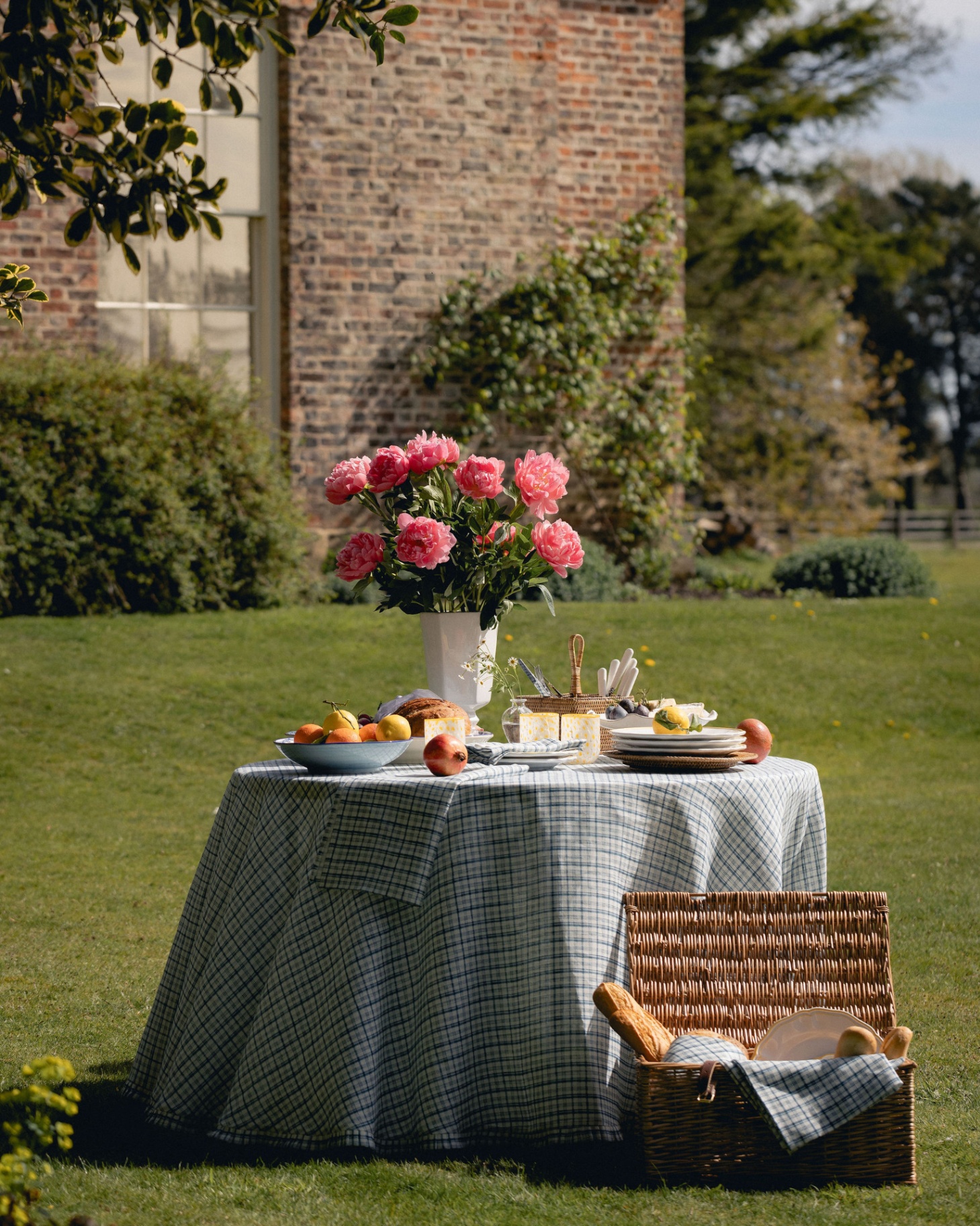 a circle table on a field covered in a blue check tablecloth, a white vase with pink flowers, bowls of food and a stack of plates and a wicker basket on the floor filled with bread