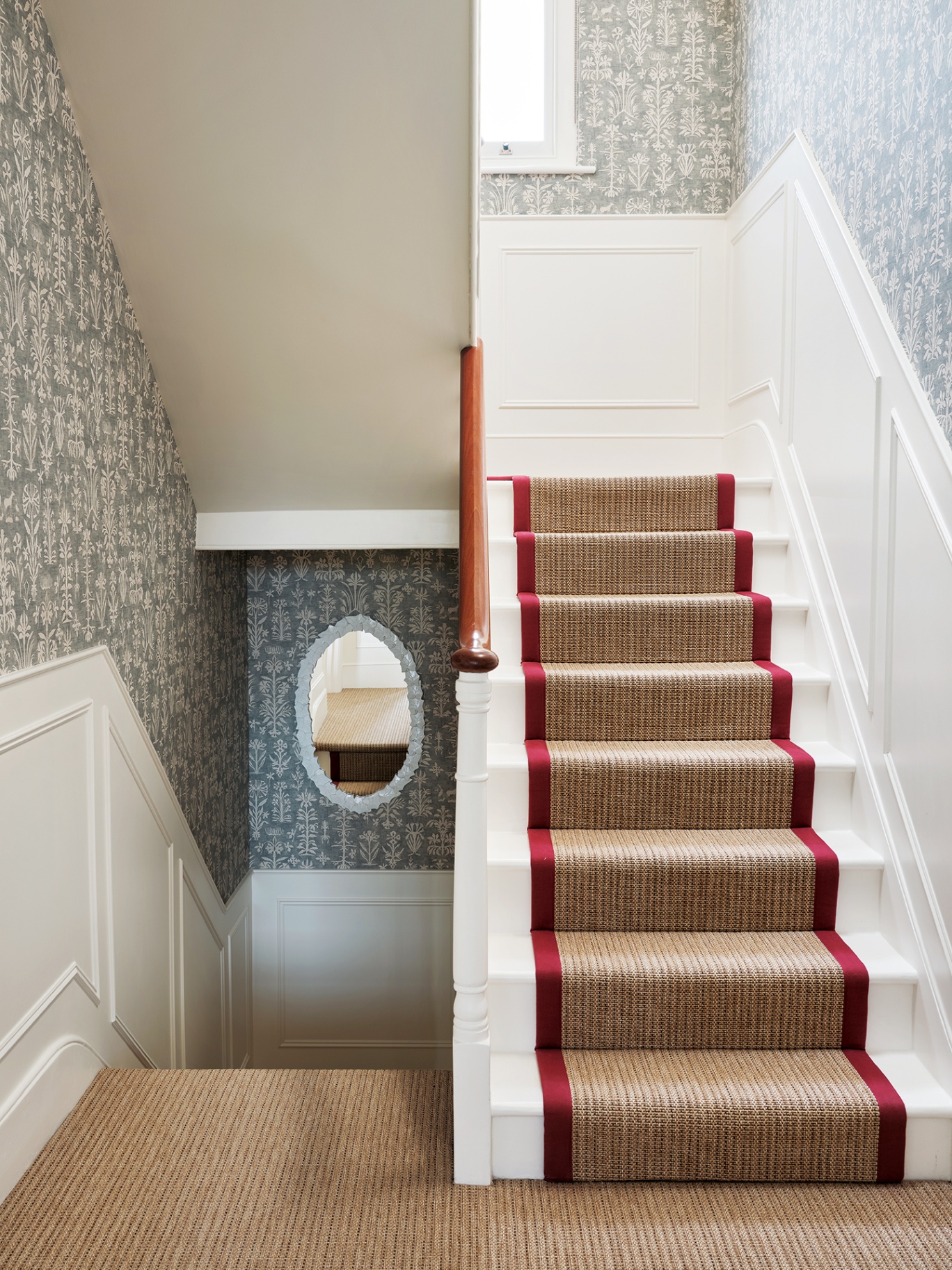 A townhouse stairway with blue wallpaper on the upper half of the walls and white panelling on the lower half. Jute carpet covers the floors and runs up the stairs, with a red border on the runner.
