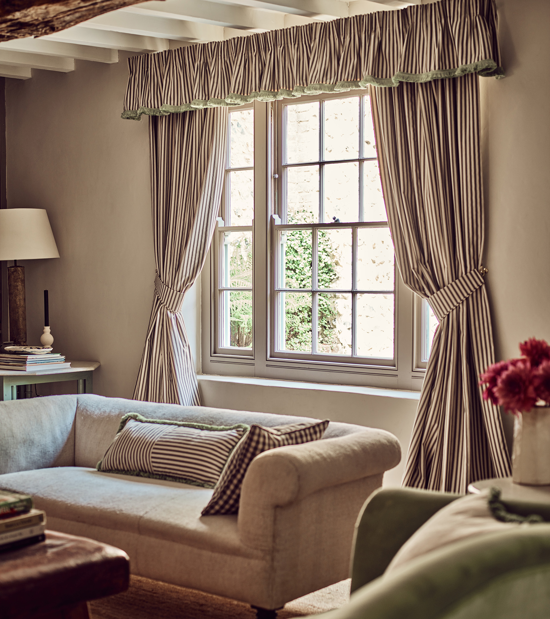 a corner of a living room with white walls and wooden beams, a large window with white and black stripe curtains with a neutral sofa infront