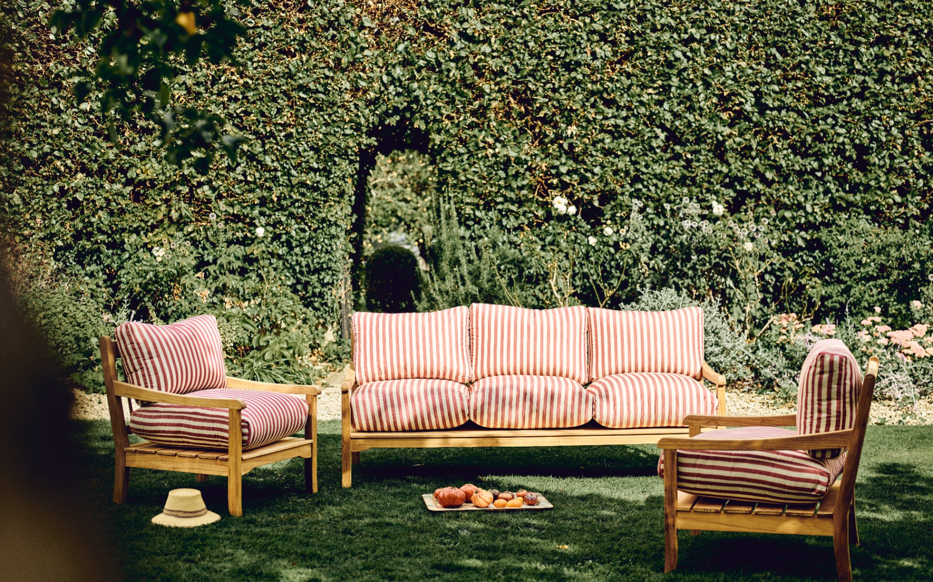 a wooden garden sofa and two armchairs with white and red striped cushions on grass with a large hedge behind it