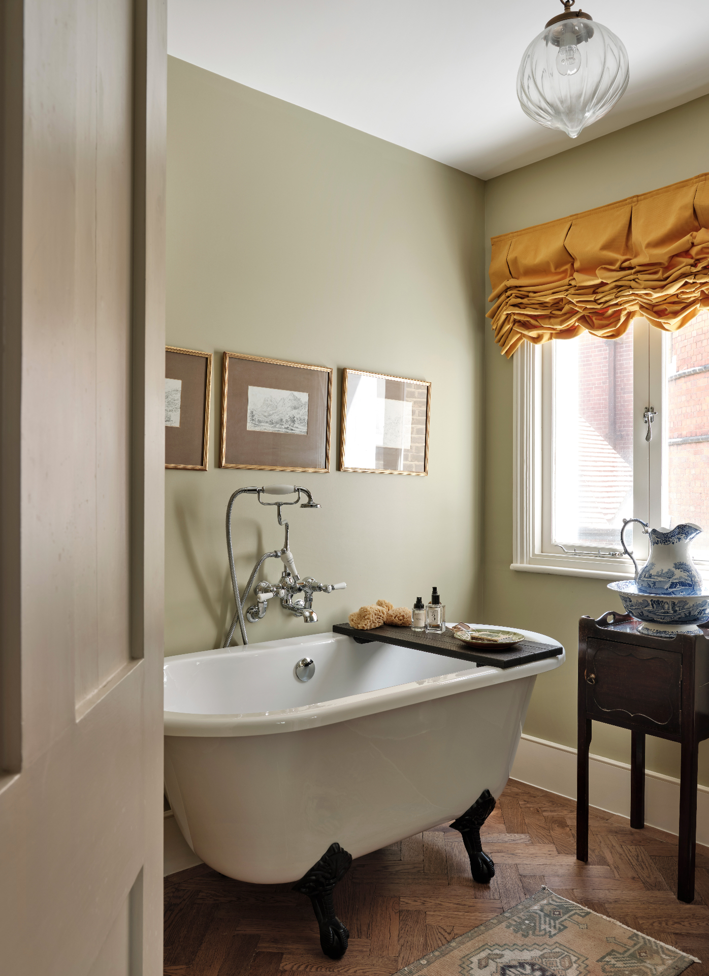 a neutral bathroom with wooden floors, a freestanding bath tub, 3 paintings on the wall above the bath and an ochre yellow blind on the window