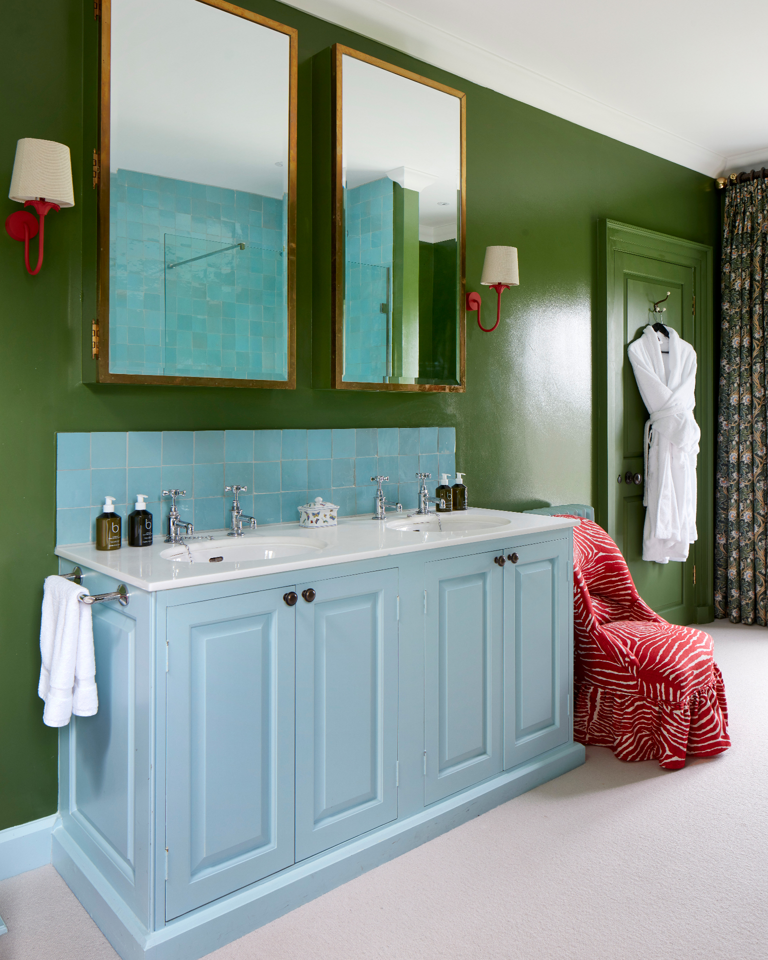 a bathroom with bright green walls, a pale blue vanity unit with double sinks, two large gold mirrors and a red and white patterned slipper chair