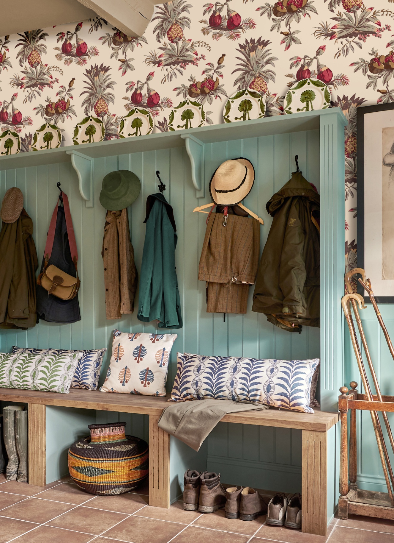a boot room with pale blue tongue and groove walls, a wooden bench with cushions, hooks with hats and coats and white and red botanical wallpaper
