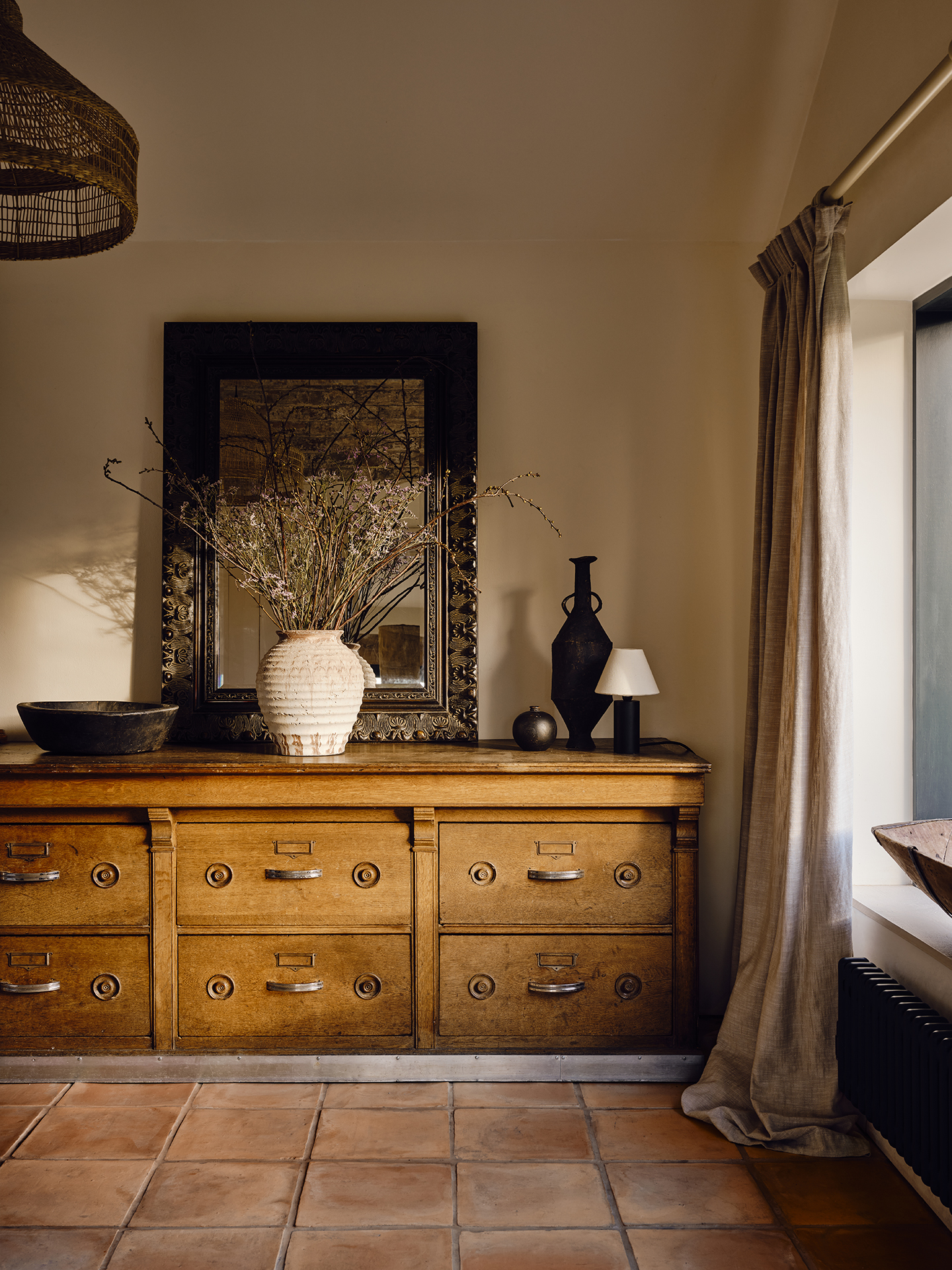 A wooden sideboard with large drawers sits against a wall, topped with a large mirror, bowl, table lamp, trinkets and a white vase with dried flowers
