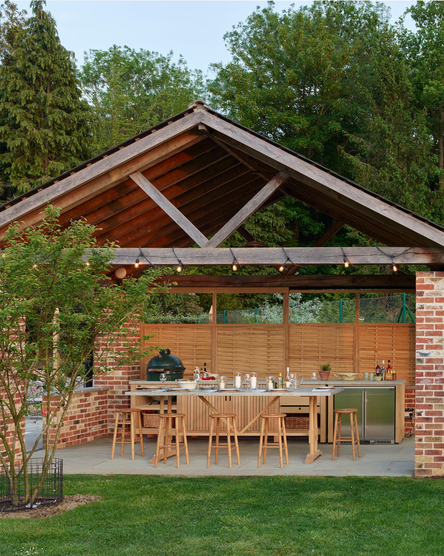 an outdoor kitchen under a brick shelter with lights wrapped around the beams, a long dining table with 5 stools, and outdoor kitchen behind the table with a big green egg bbq and a tree to the left