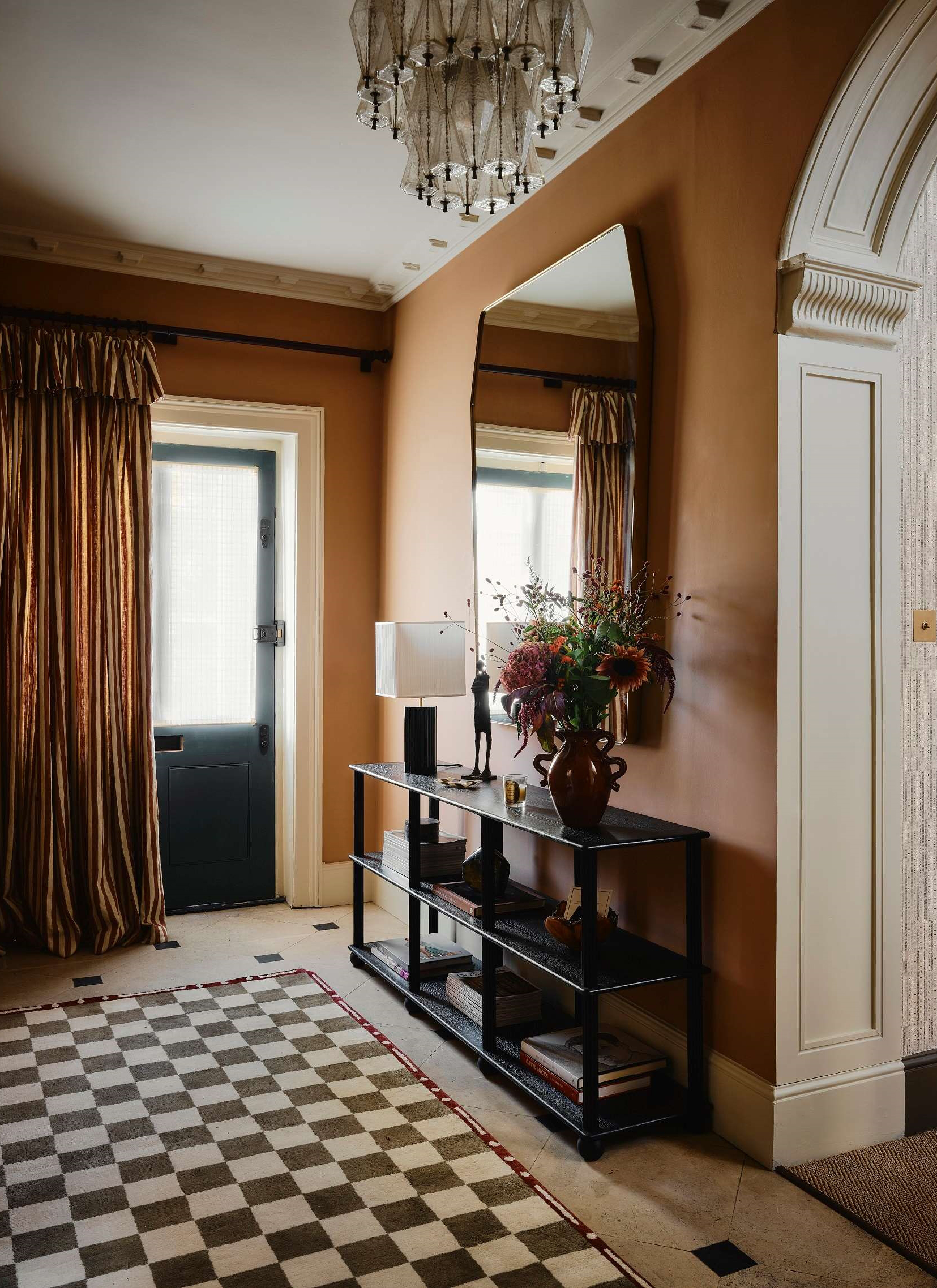 A hallway with caramel wall,s a stripe door curtain, a large mirror and a green and white checkerboard rug