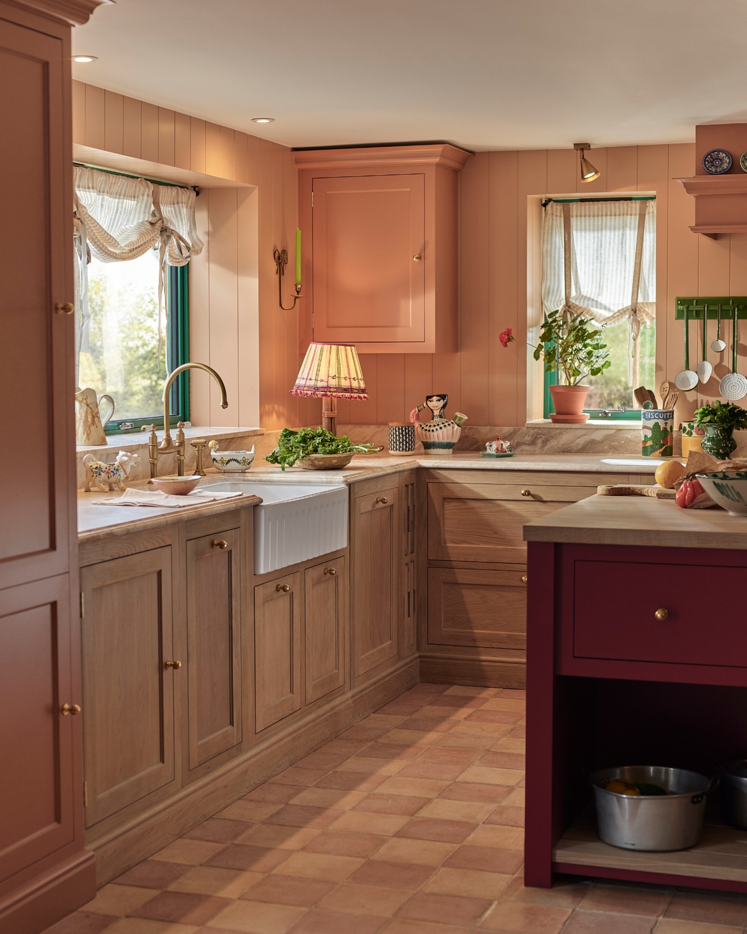A kitchen with terracotta checkered floor tailes, a red kitchen island, wooden cabinetry at the bottom and terracotta pink cabinets higher up the walls