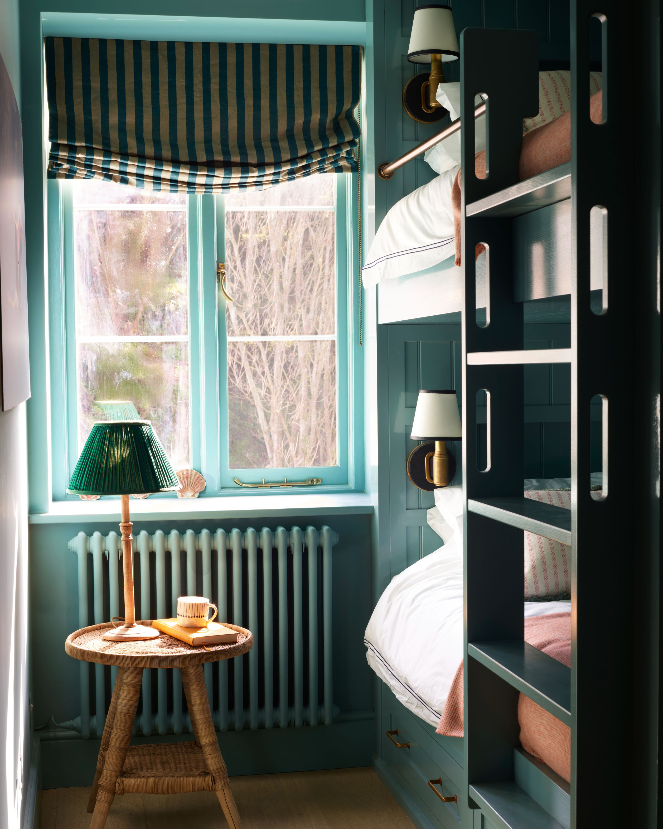a dark teal bedroom with stacked built in bunk beds, a window with teal and cream striped blinds and a rattan table with a wood and teal green pleated lamp on top