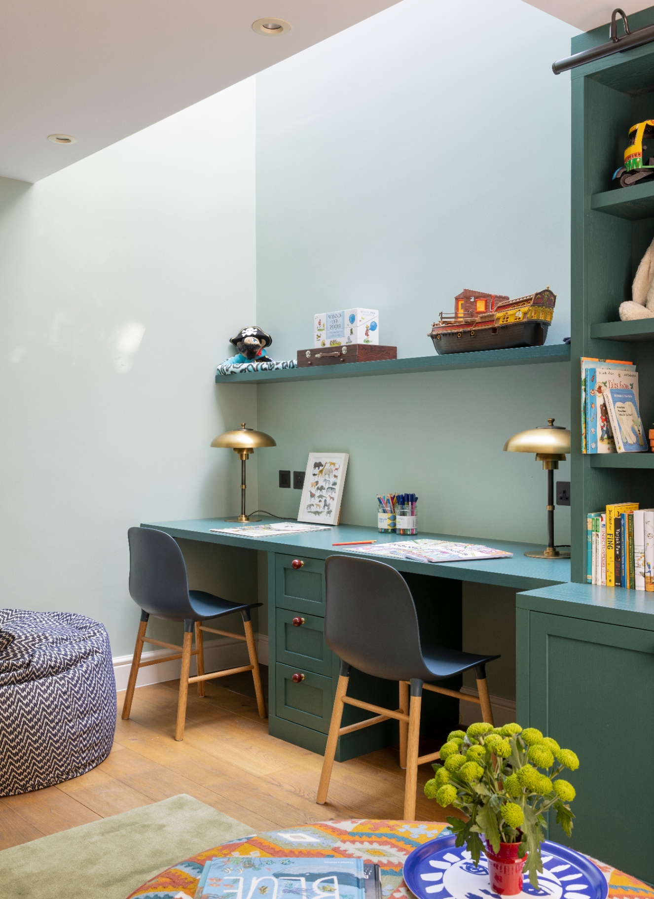 Light-filled dual workspace under a skylight with a long built-in teal desk and shelving, two modern desk chairs, and brass task lamps