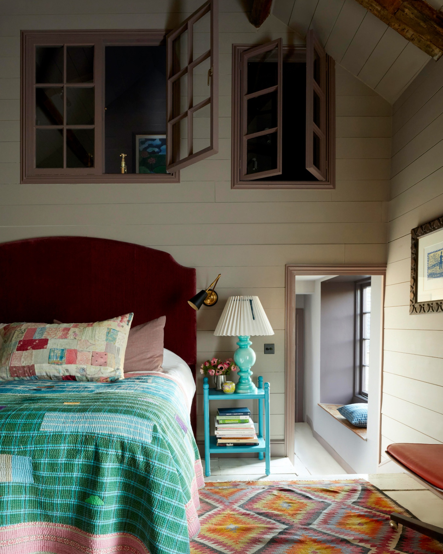 a cottage bedroom with cream tongue and groove panelled walls with two internal windows above the bed, a bed with a dark red velvet headboard and bright patchwork bedding, a bright teal blue bedside table with a turqoise lamp with a pleated white shade