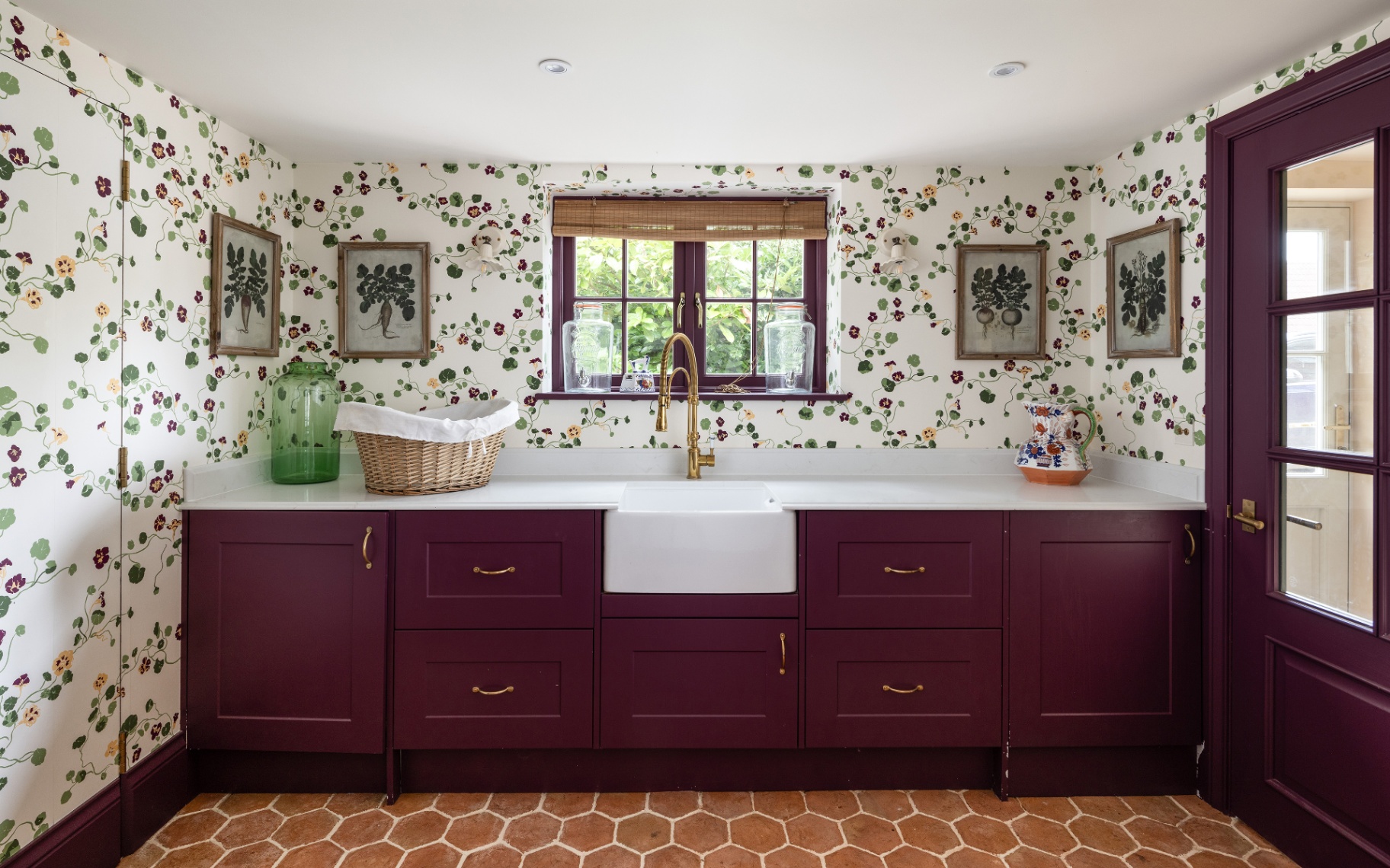 a utility room with white and green leafy botanical wallpaper, aubergine purple cabinetry, botanical prints in frames on the wall, brick flooring, a wicker laundry basket on the worktop
