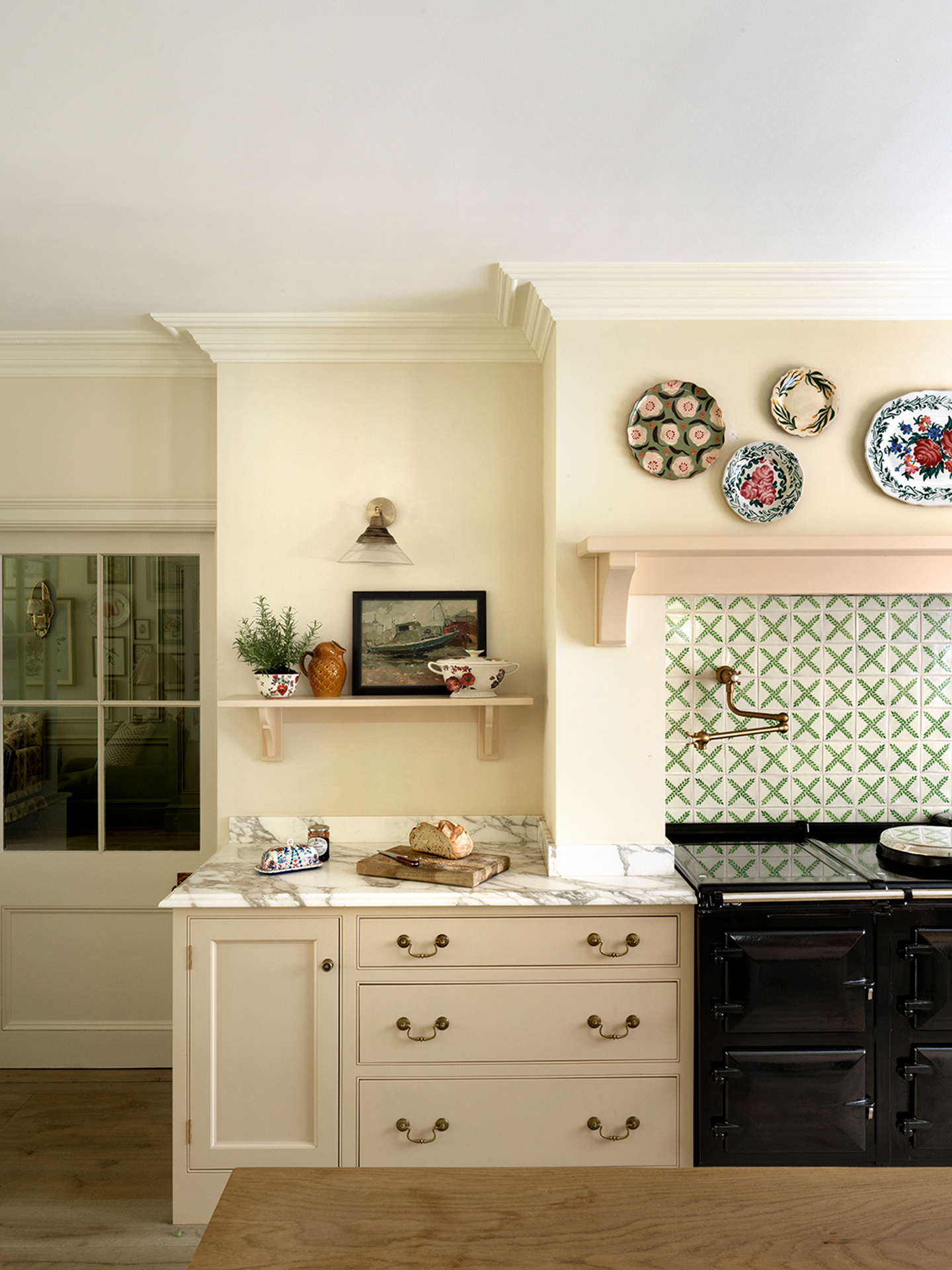 A close up of a kitchen with neutral lower cabinets topped with marble, a black aga with a green-and-white tiled backsplash and a corbel and mantel above, decorative wall plates, and an open shelf topped with ceramics and a painting